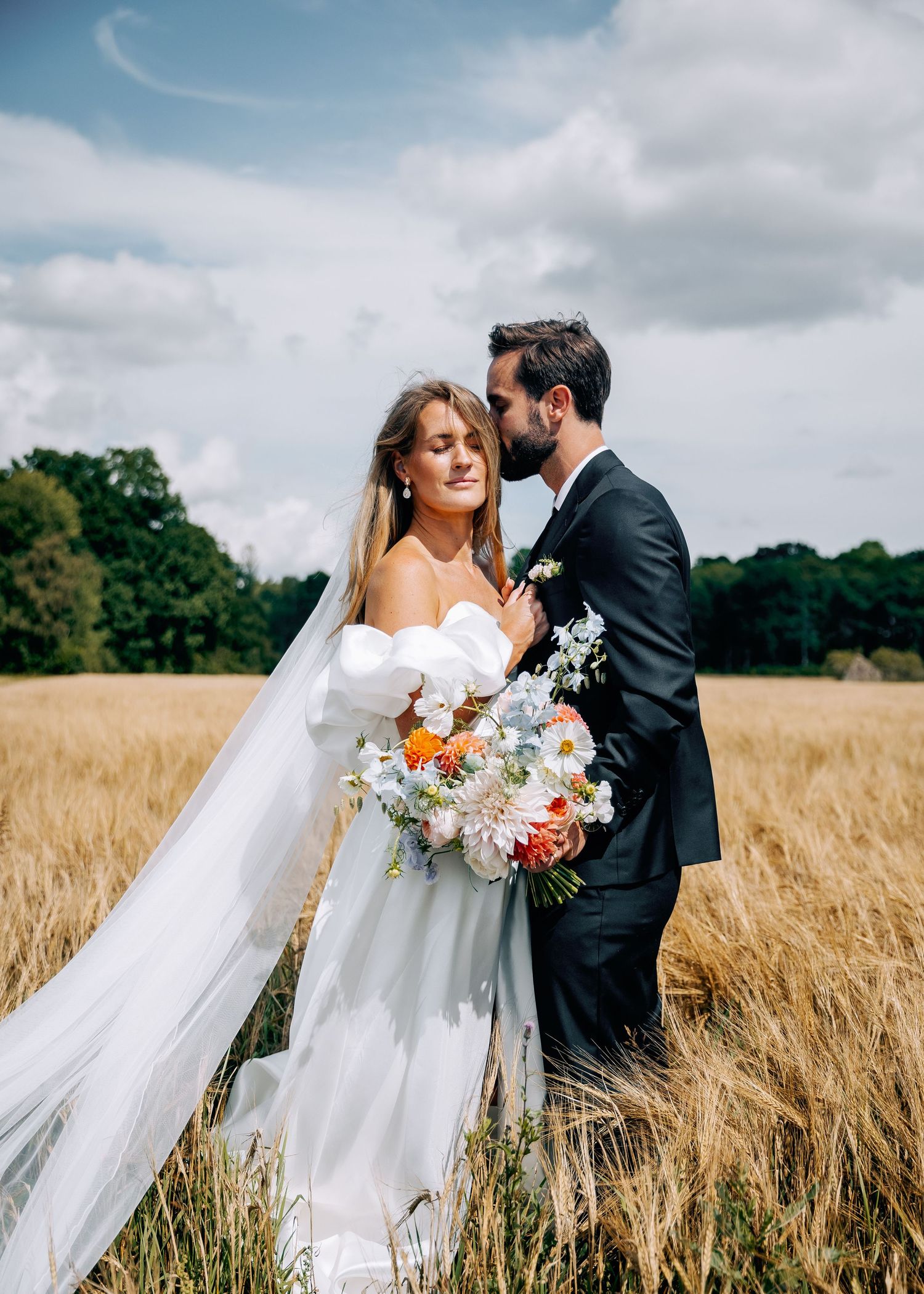 Romantic wedding portrait in wheat field with flowing veil and floral bouquet.