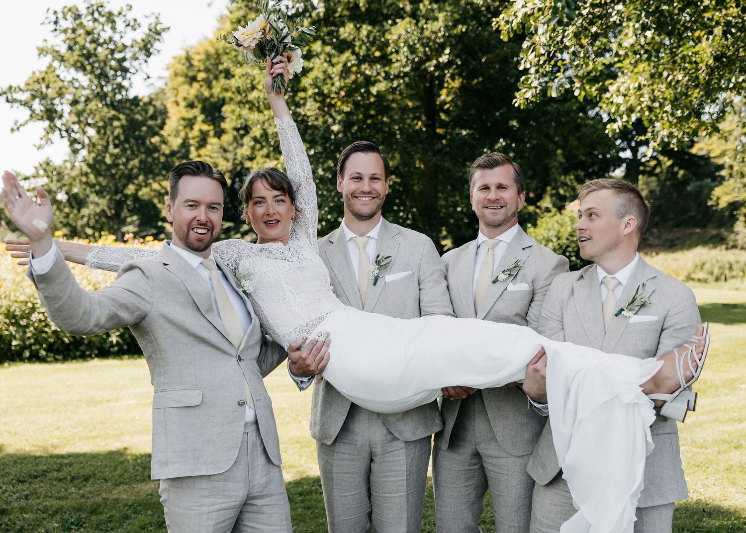 Groomsmen in light gray suits pose playfully outdoors under trees.