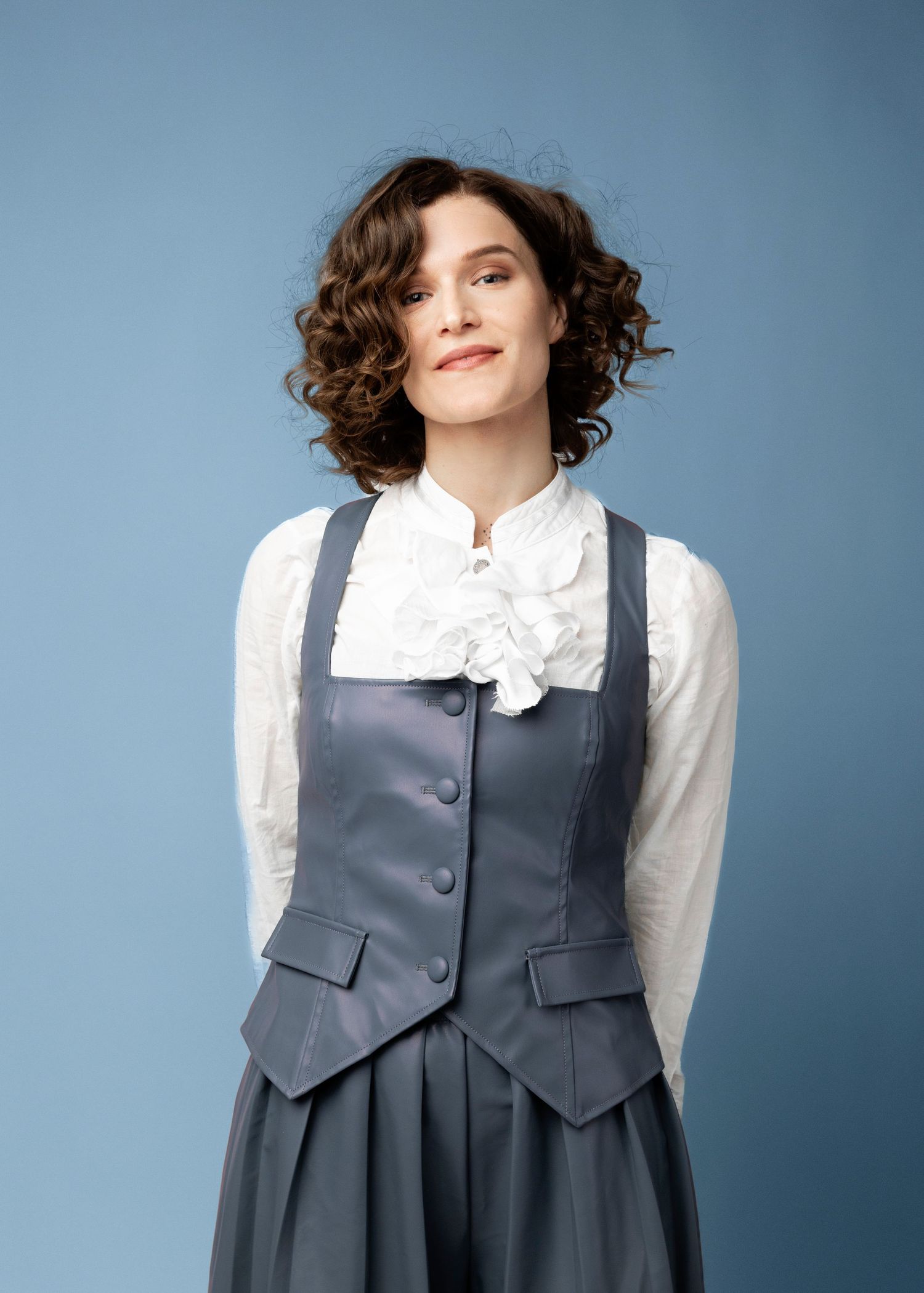 Fashion photo of model wearing grey vest and white blouse against blue background.