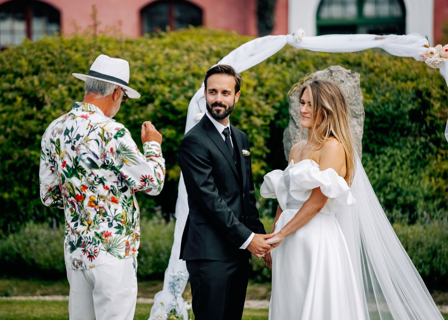 Wedding ceremony moments captured against pink building with white draped arch and greenery.