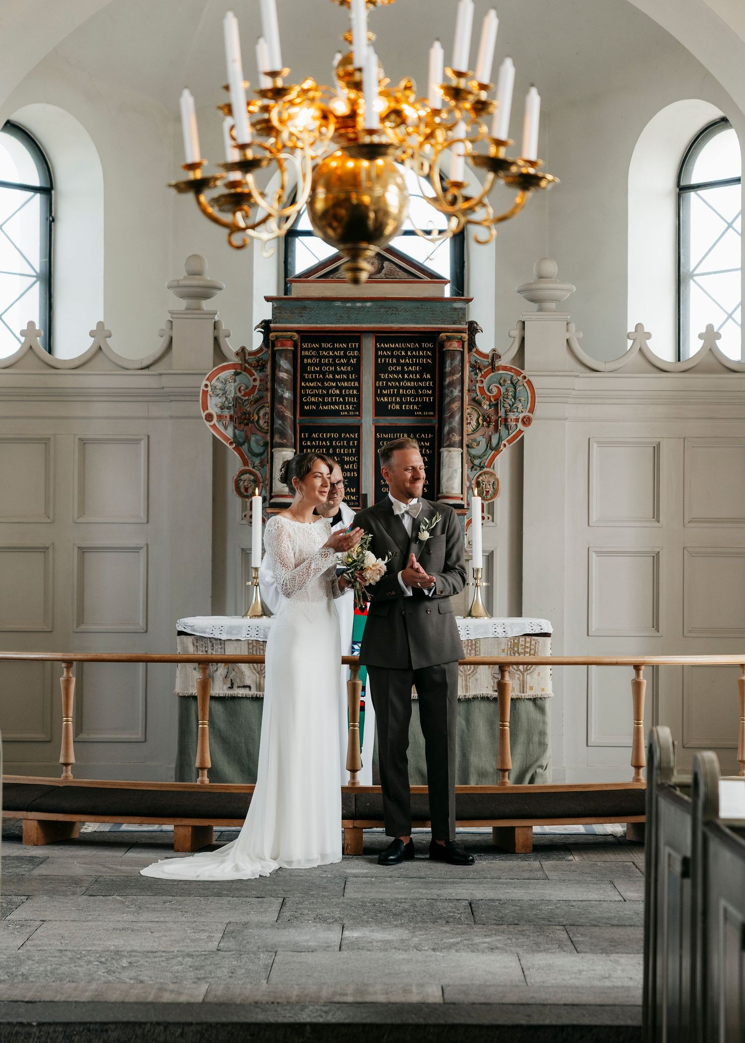 Church wedding ceremony under ornate chandelier with white walls and stone floor.