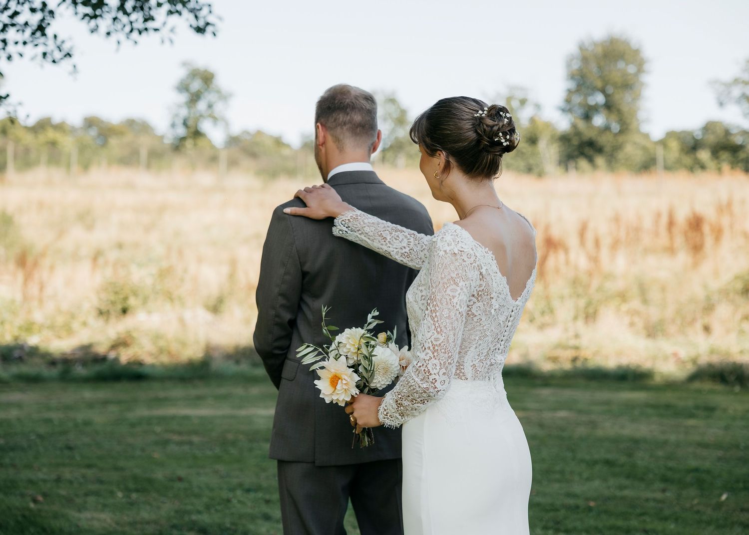 A couple in wedding attire embrace while standing in a sunlit field with wildflowers.