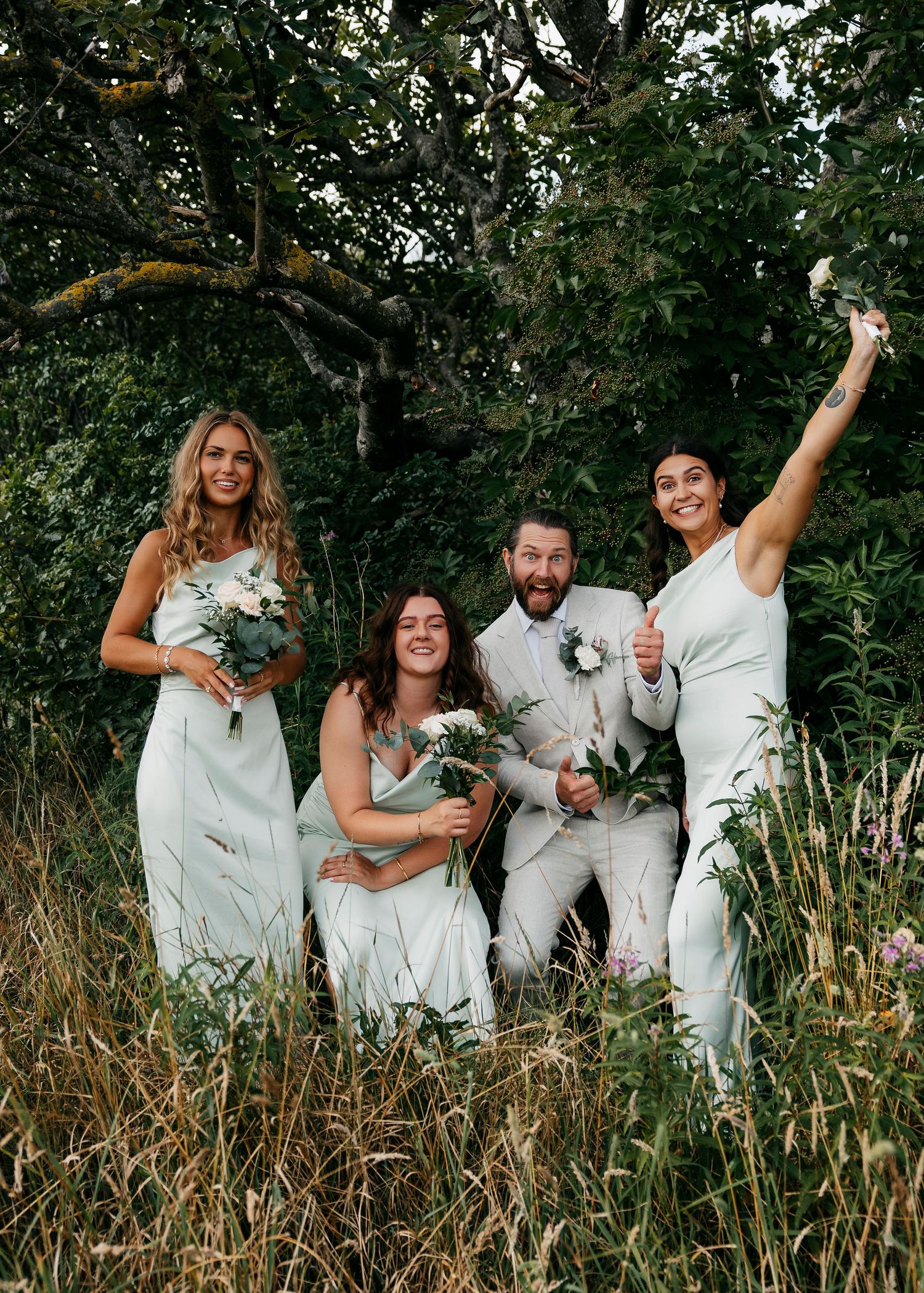 Wedding party in white outfits poses playfully in a field with tall grass and trees.