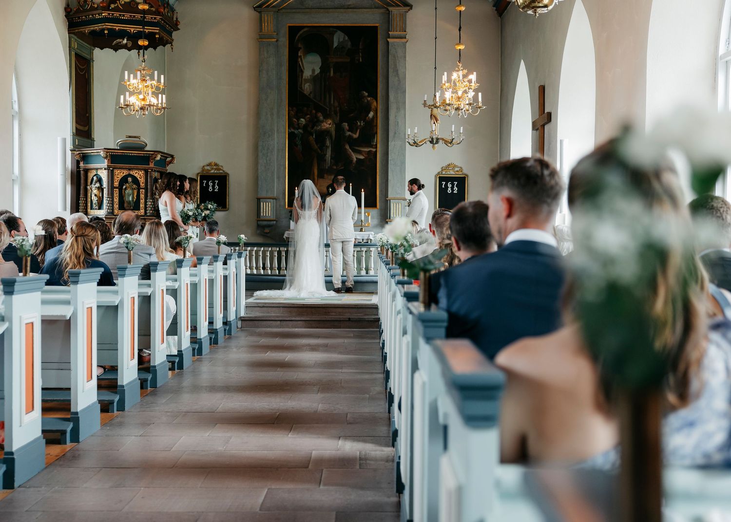 Long church aisle with ornate chandeliers and white pews lined with decorative floral arrangements.
