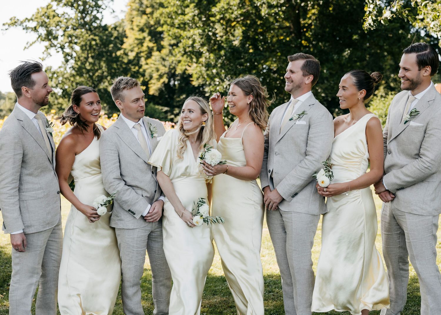 Wedding party lines up outdoors wearing coordinating white dresses and light gray suits.