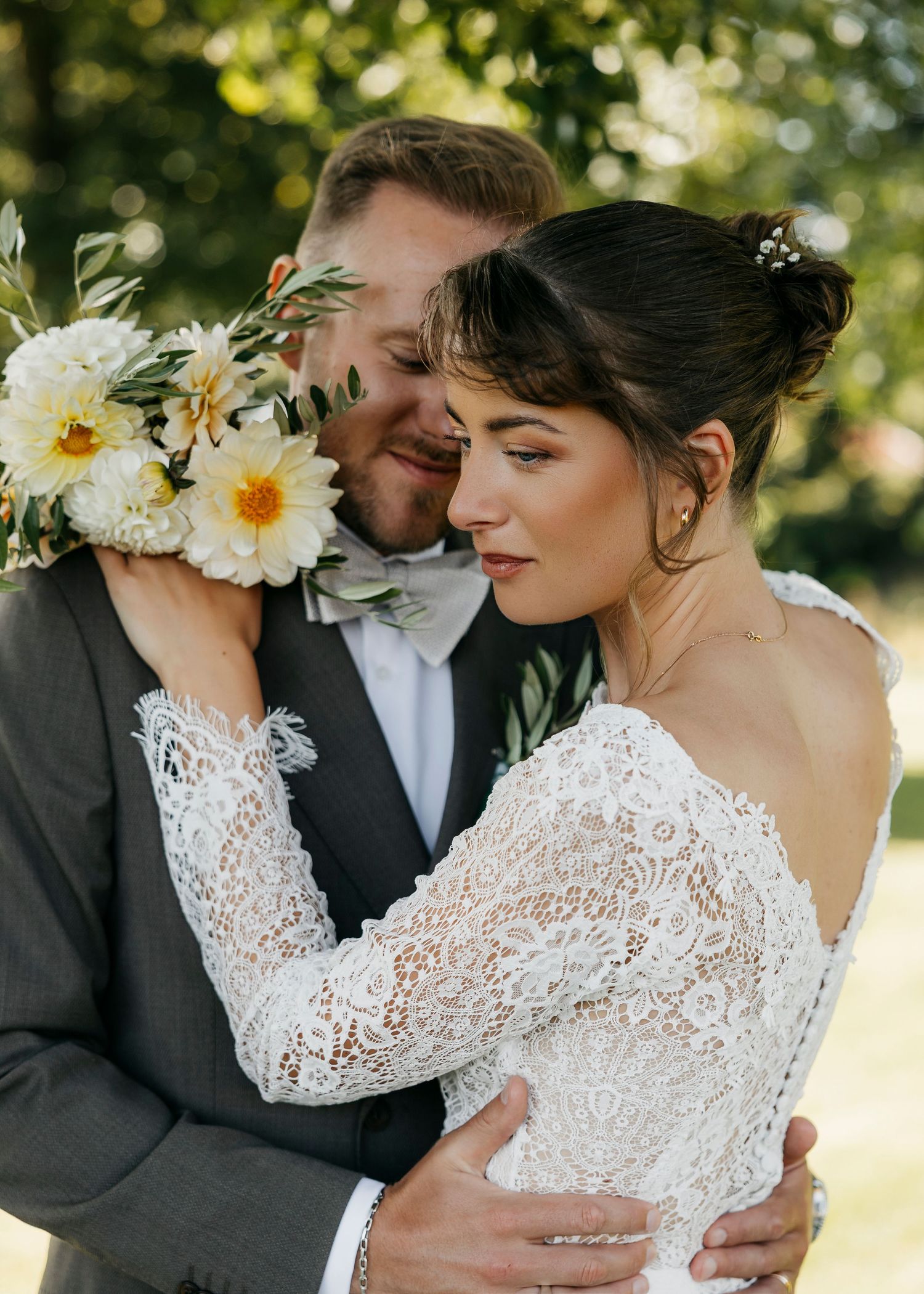 Romantic wedding couple shares intimate moment among sunlit trees with white daisies.