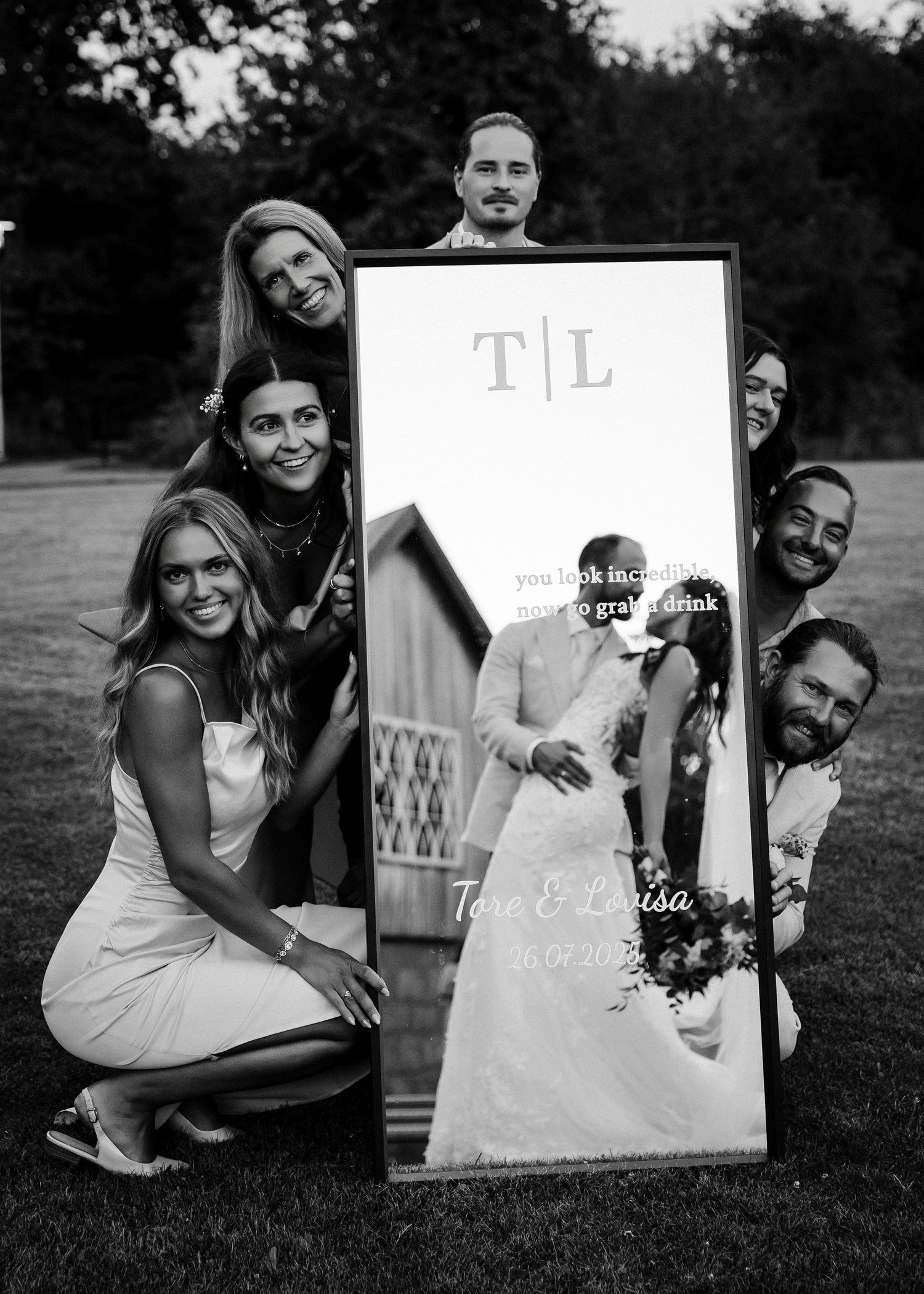 A group poses outdoors with a large framed wedding photo display showing a bride and groom.