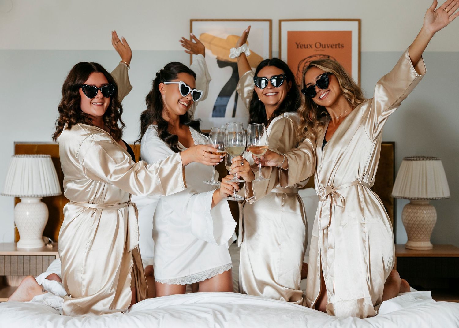 Group celebrating in white robes on hotel bed with champagne and confetti.