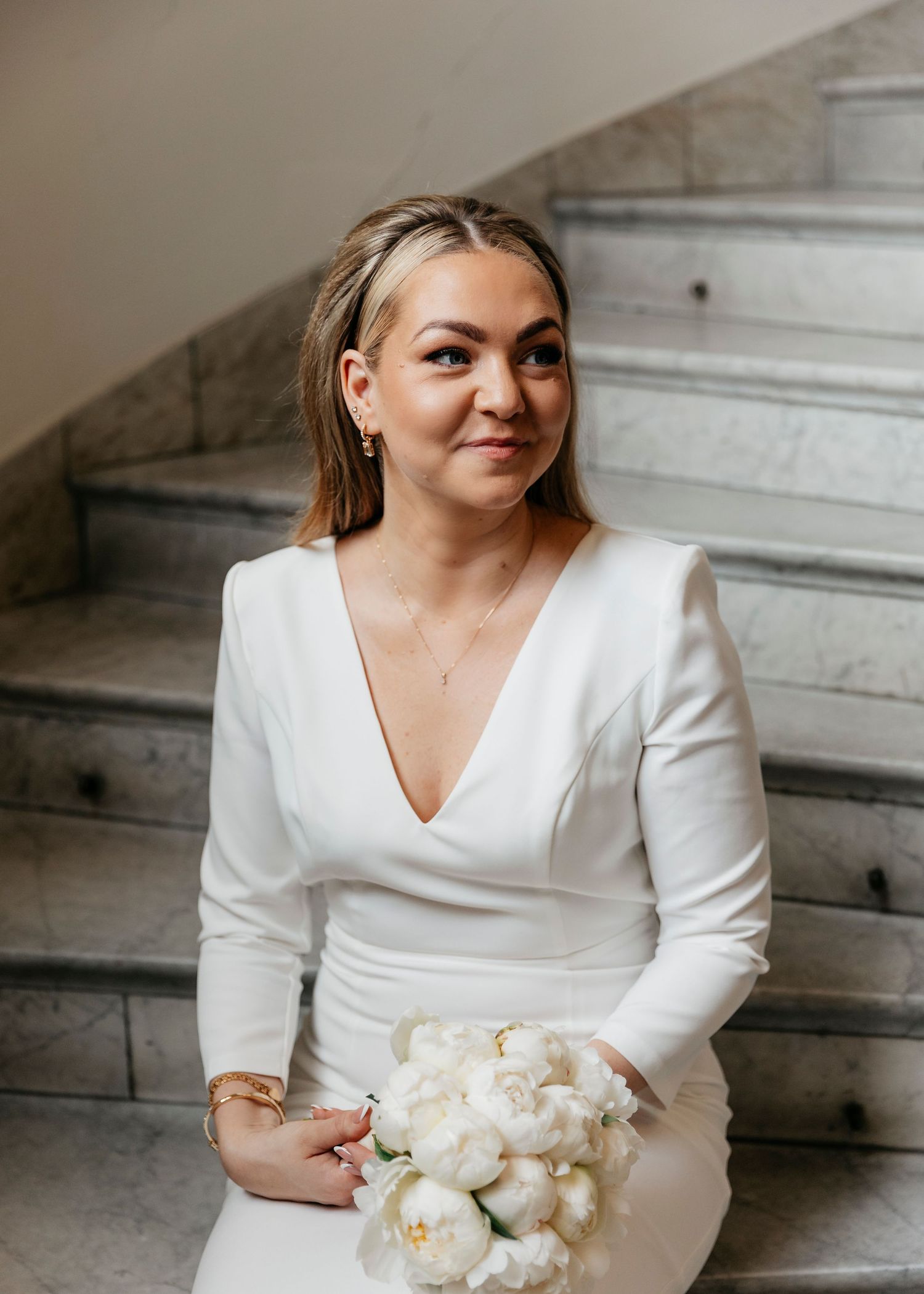 A person in a white long-sleeved dress holds a bouquet of white flowers on marble stairs.