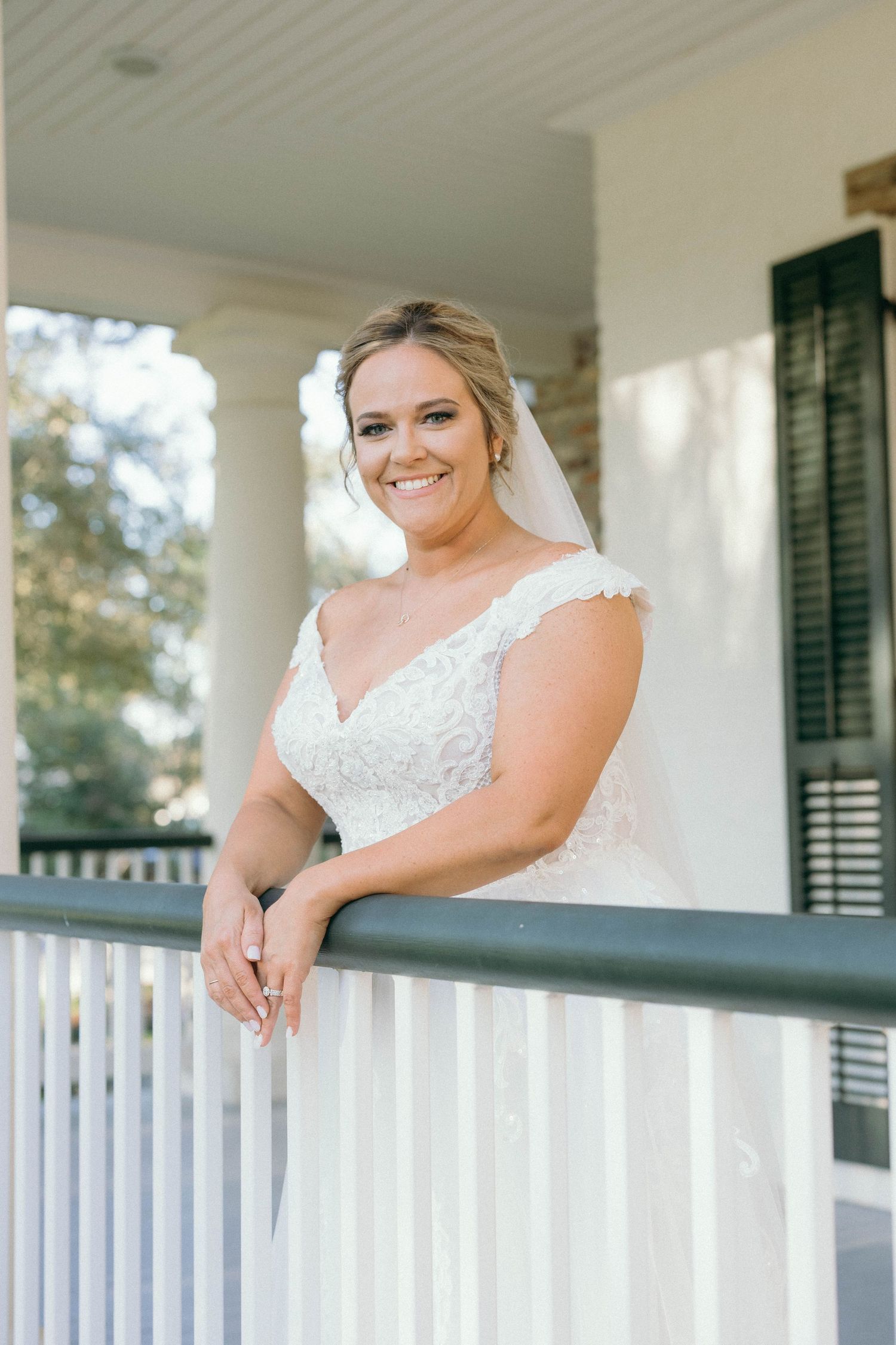 A bride in a white lace dress leans against a white porch railing on a sunny day.