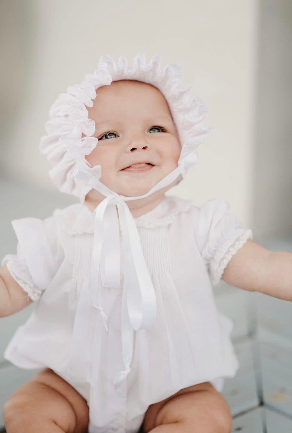 A baby wearing a white bonnet and dress smiles while looking up.