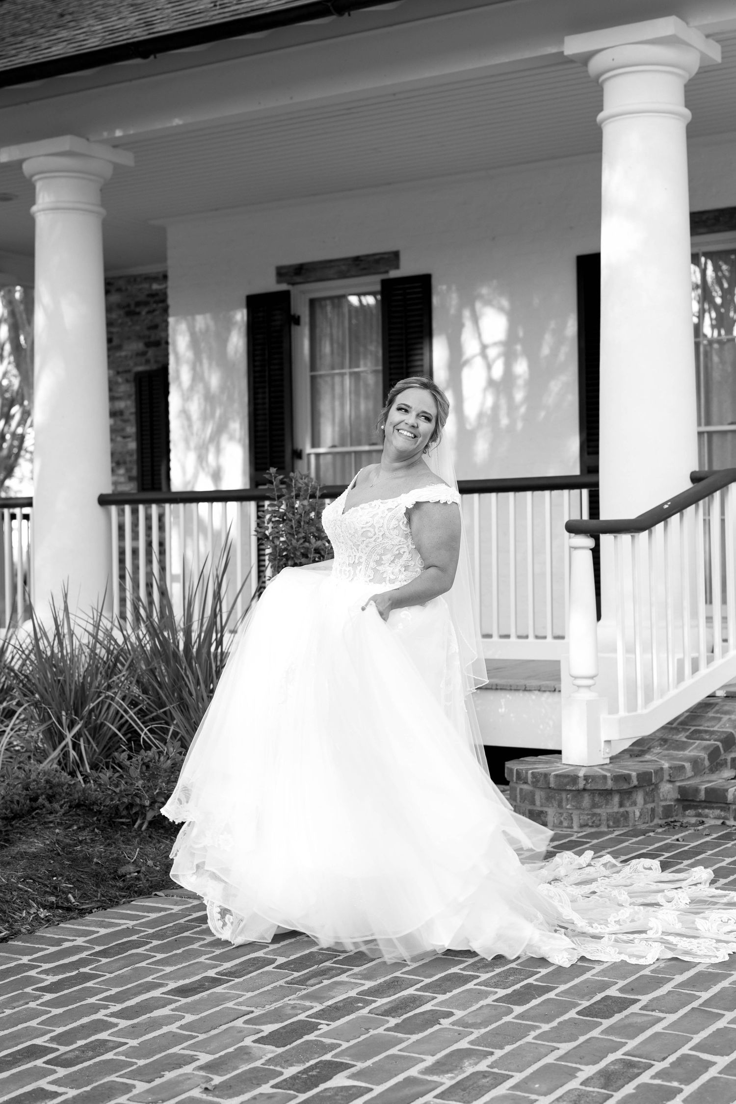 A bride in an elegant off-shoulder white wedding gown poses on a Southern-style porch with classic white columns.