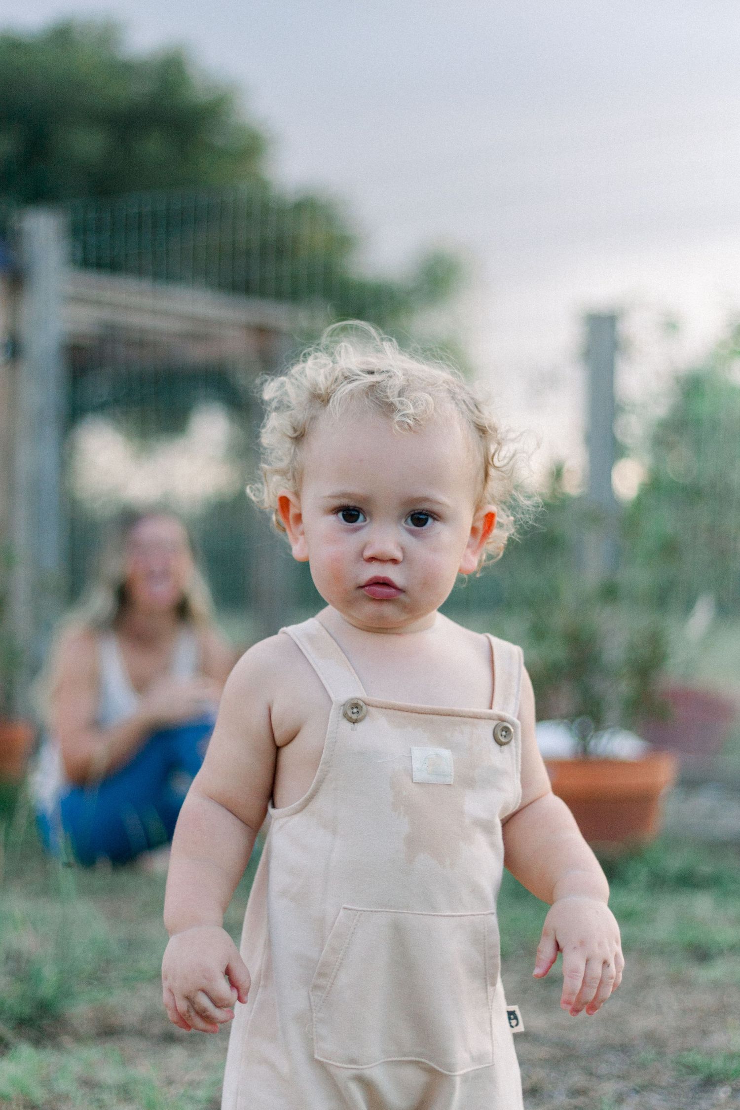 A toddler wearing beige overalls stands in a garden setting during golden hour.