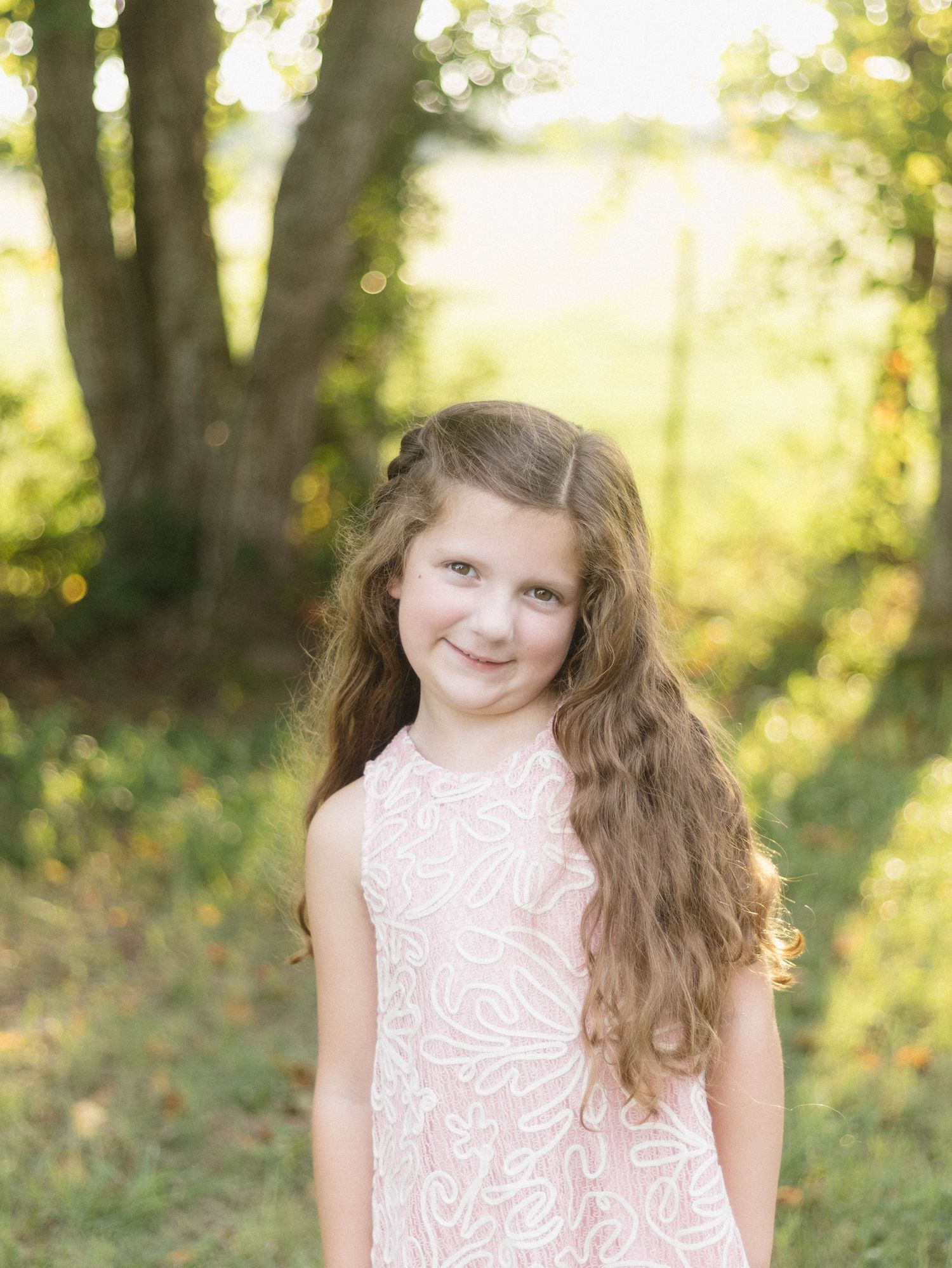 Young person in pink lace dress poses outdoors with natural bokeh background.