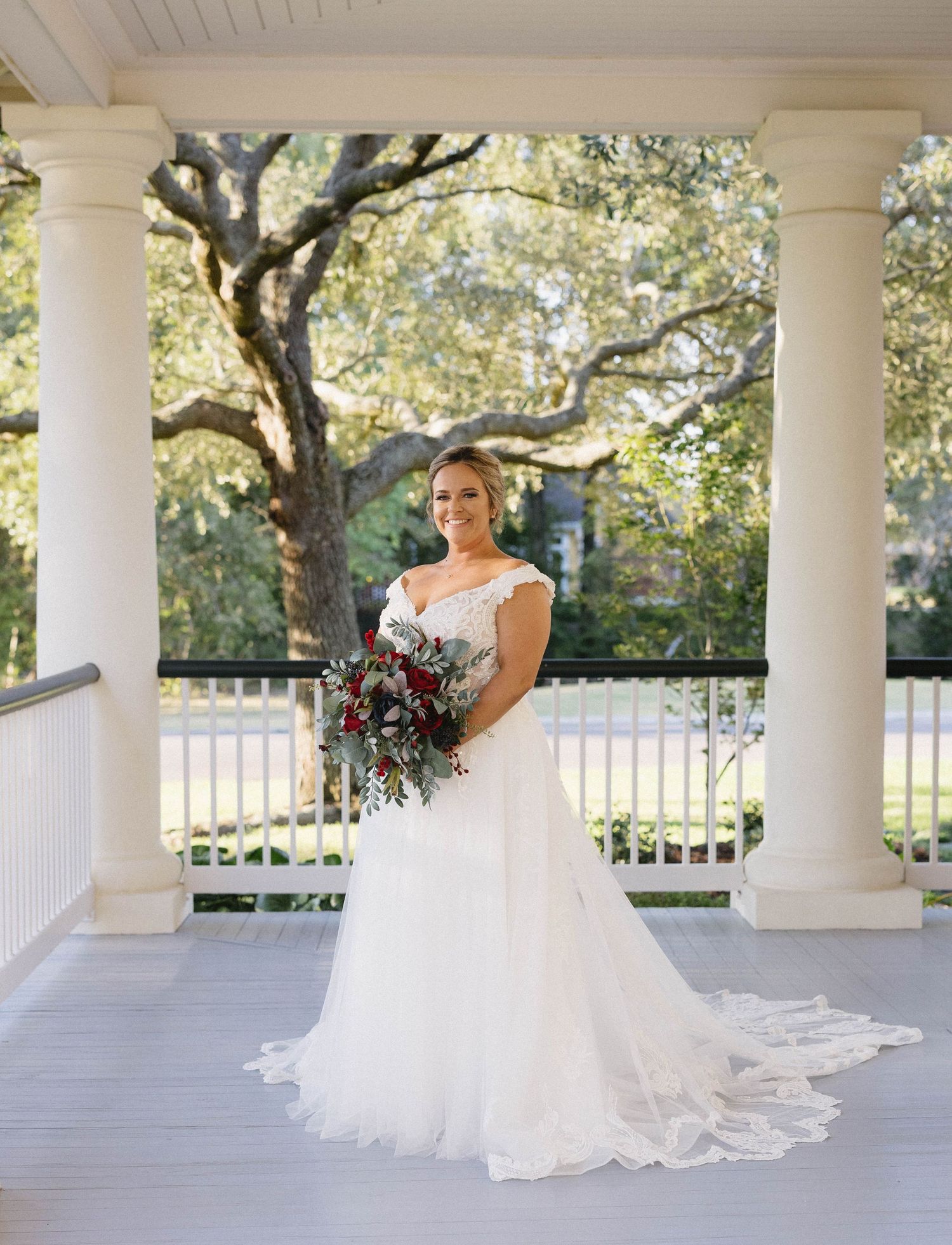 A bride in a flowing white gown poses gracefully on a white columned porch with trees visible in the background.