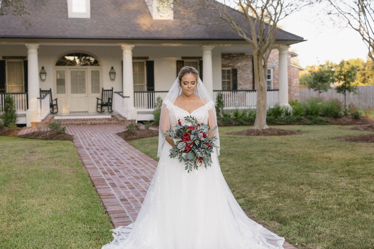 A bride in a white lace gown holds a vibrant bouquet while standing on a brick path leading to a craftsman-style house.