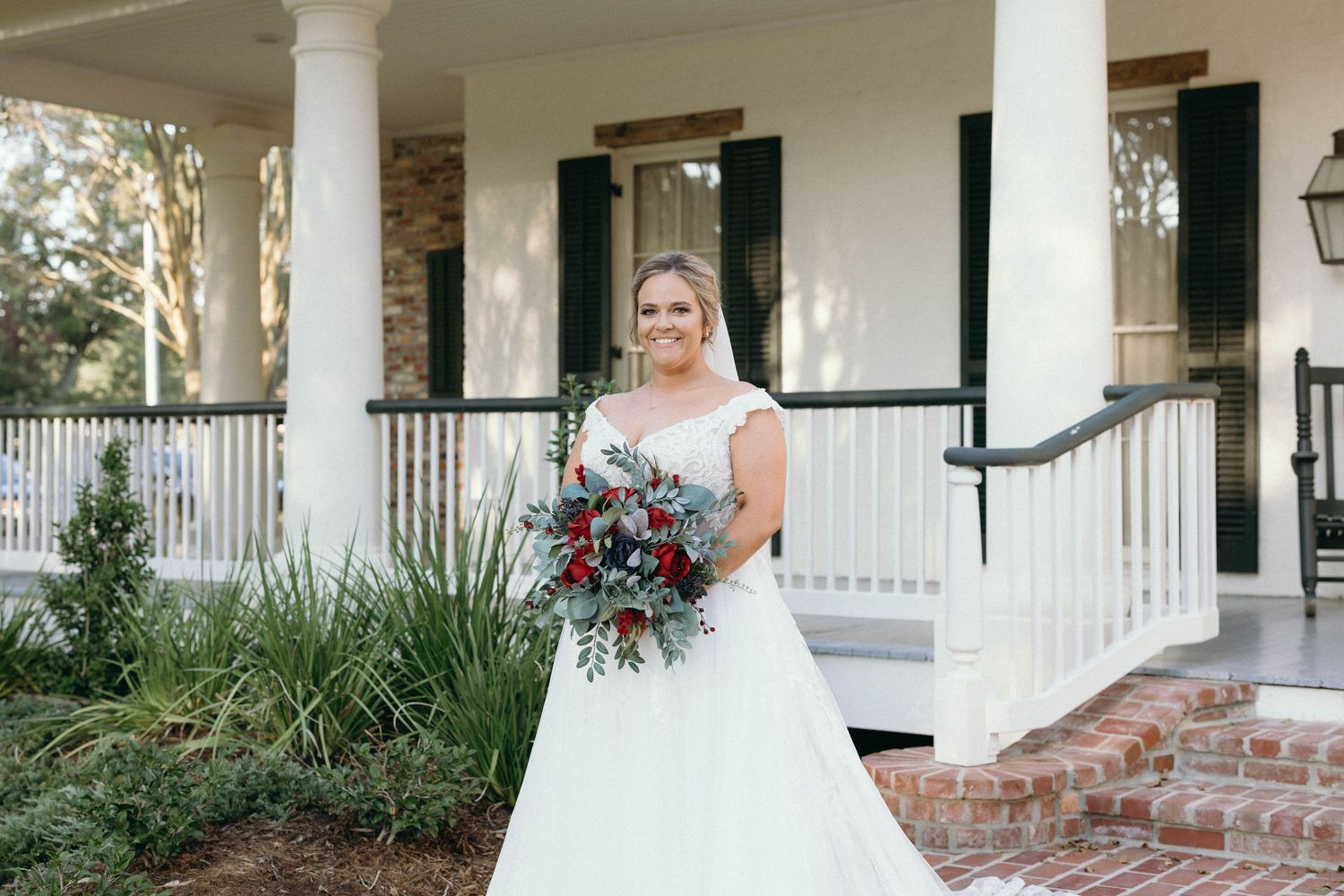 A bride in a white wedding dress poses on a charming Southern style porch with white columns and brick steps.