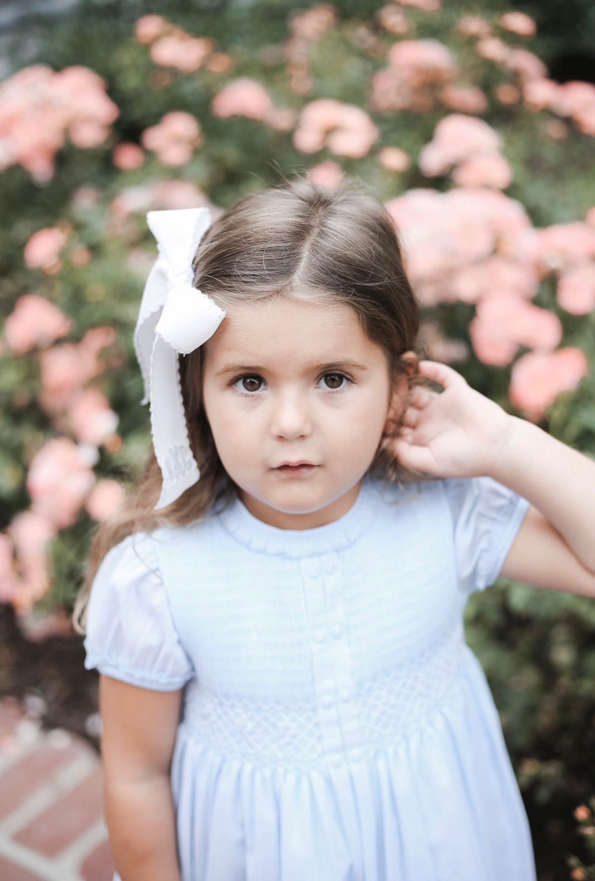 A child in a light blue dress with white bow poses against pink flowers in a garden.