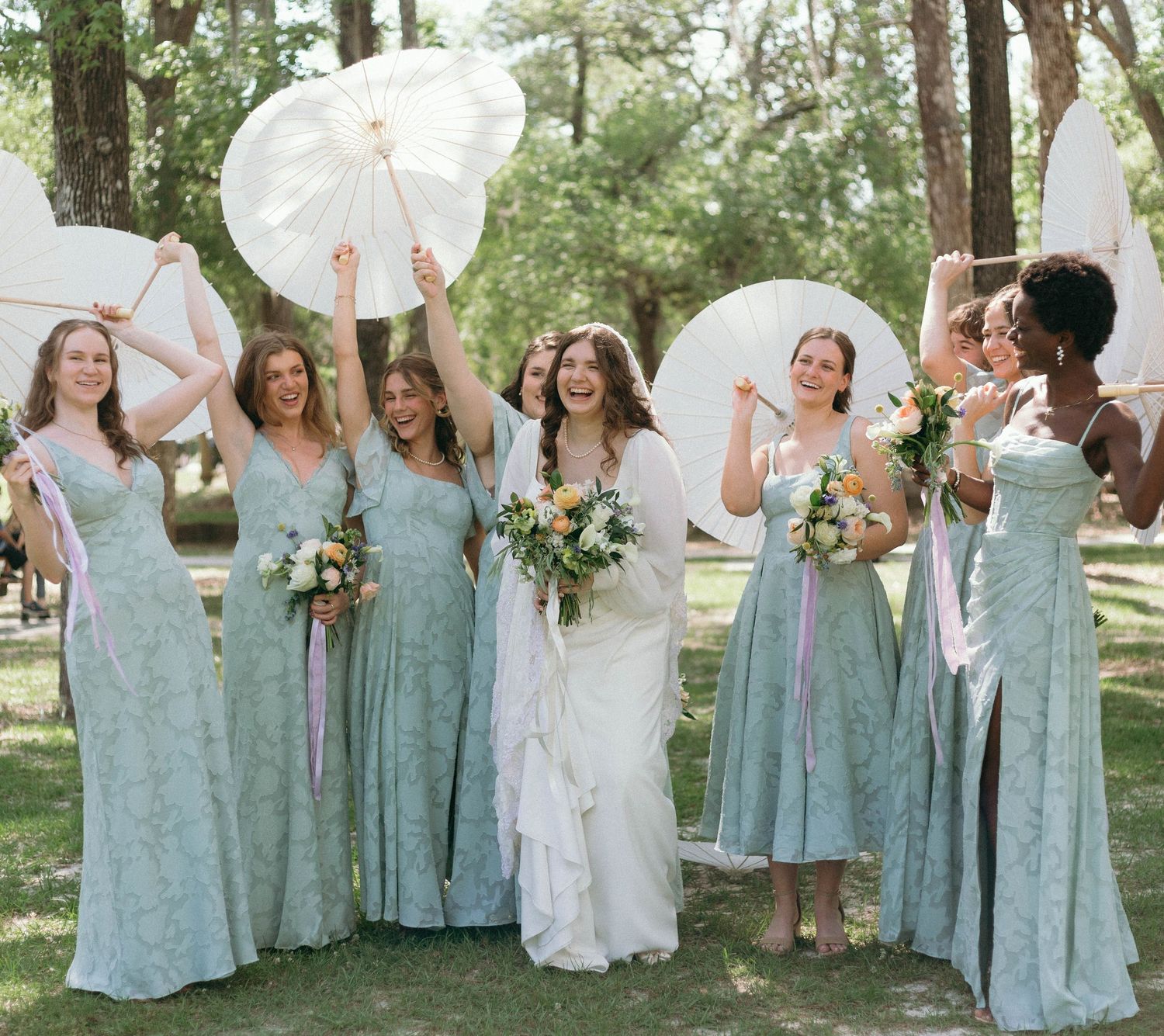 Wedding party with bridesmaids in mint green dresses holding white parasols in a garden setting.