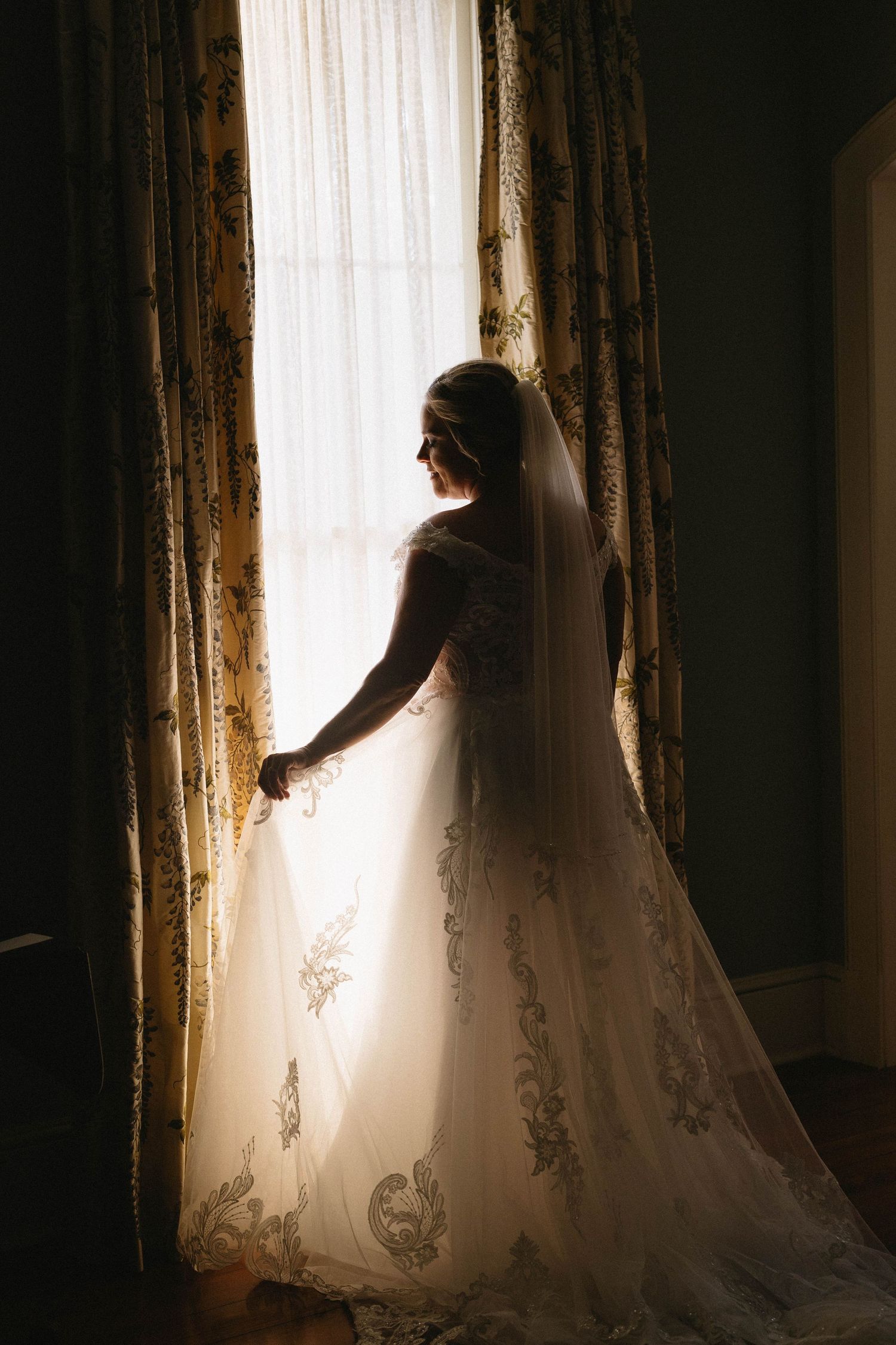 Silhouette of a bride in a flowing wedding dress standing by a sunlit window with elegant curtains.