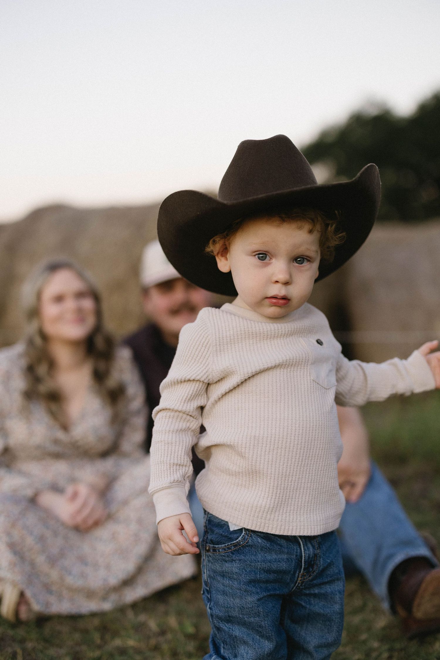 A toddler in a beige sweater and brown cowboy hat stands in a field at sunset.