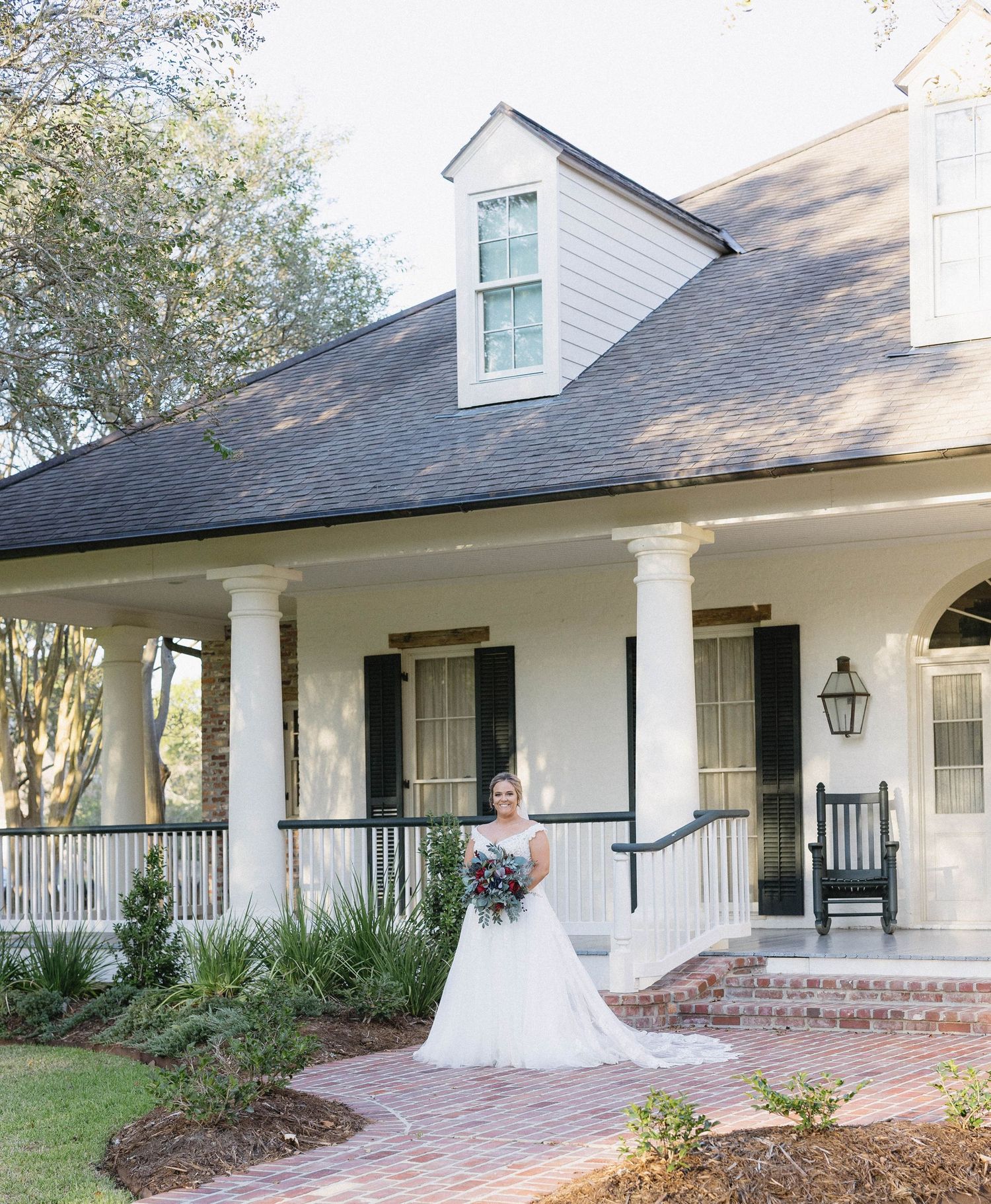 A charming white Southern-style home with a wrap-around porch, columns and dormer window sits amid landscaped grounds.