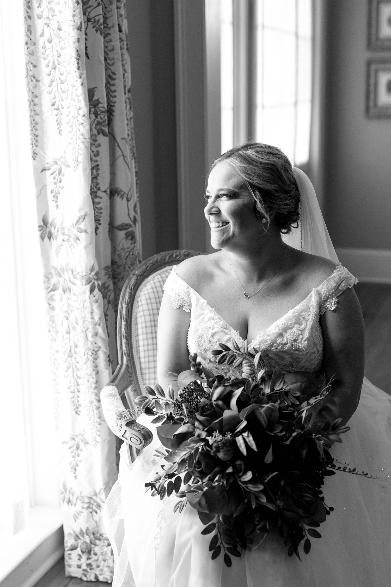 A bride in a lace wedding dress sits by a window holding a lush bouquet in black and white photography.