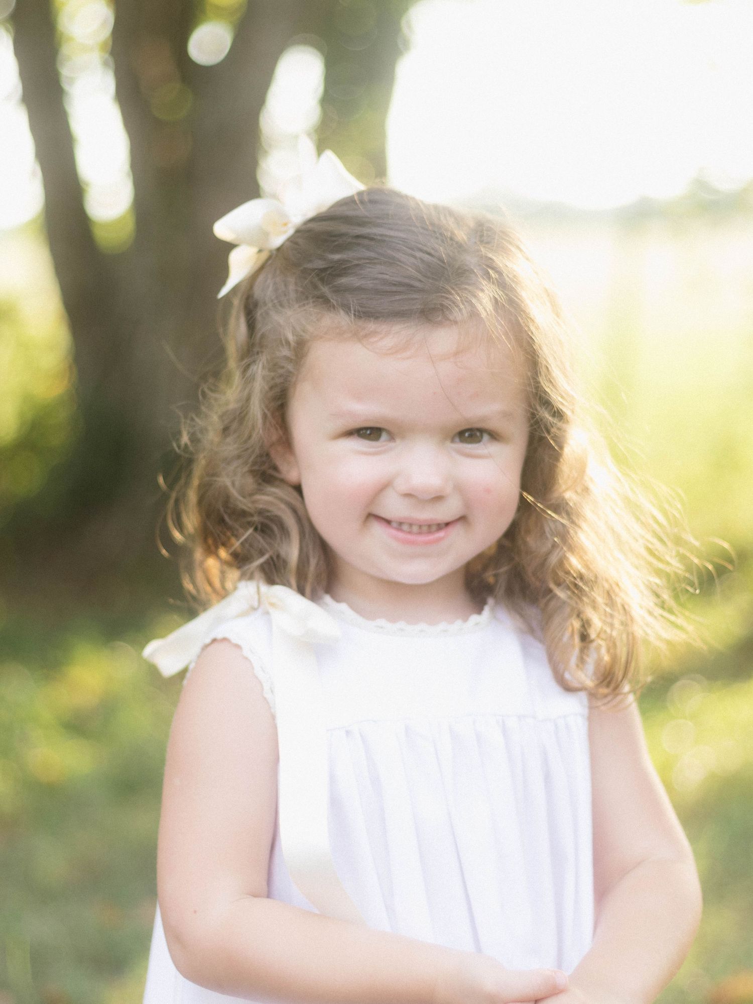 A young child in a white dress poses outdoors in golden sunlight.