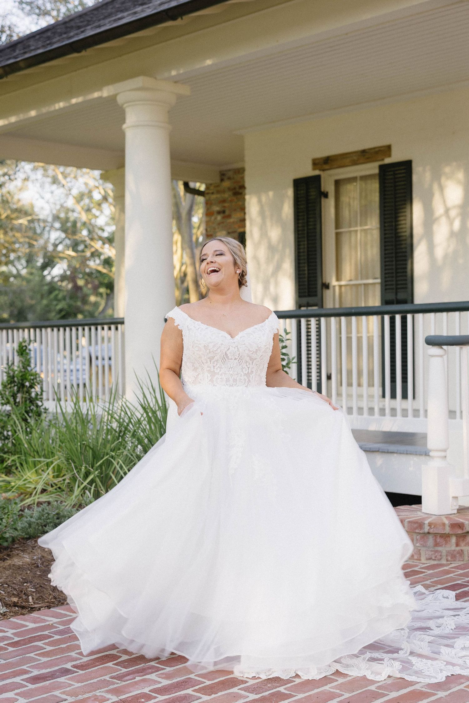 A flowing white wedding dress with delicate lace details cascades across a brick porch of a classic southern home.