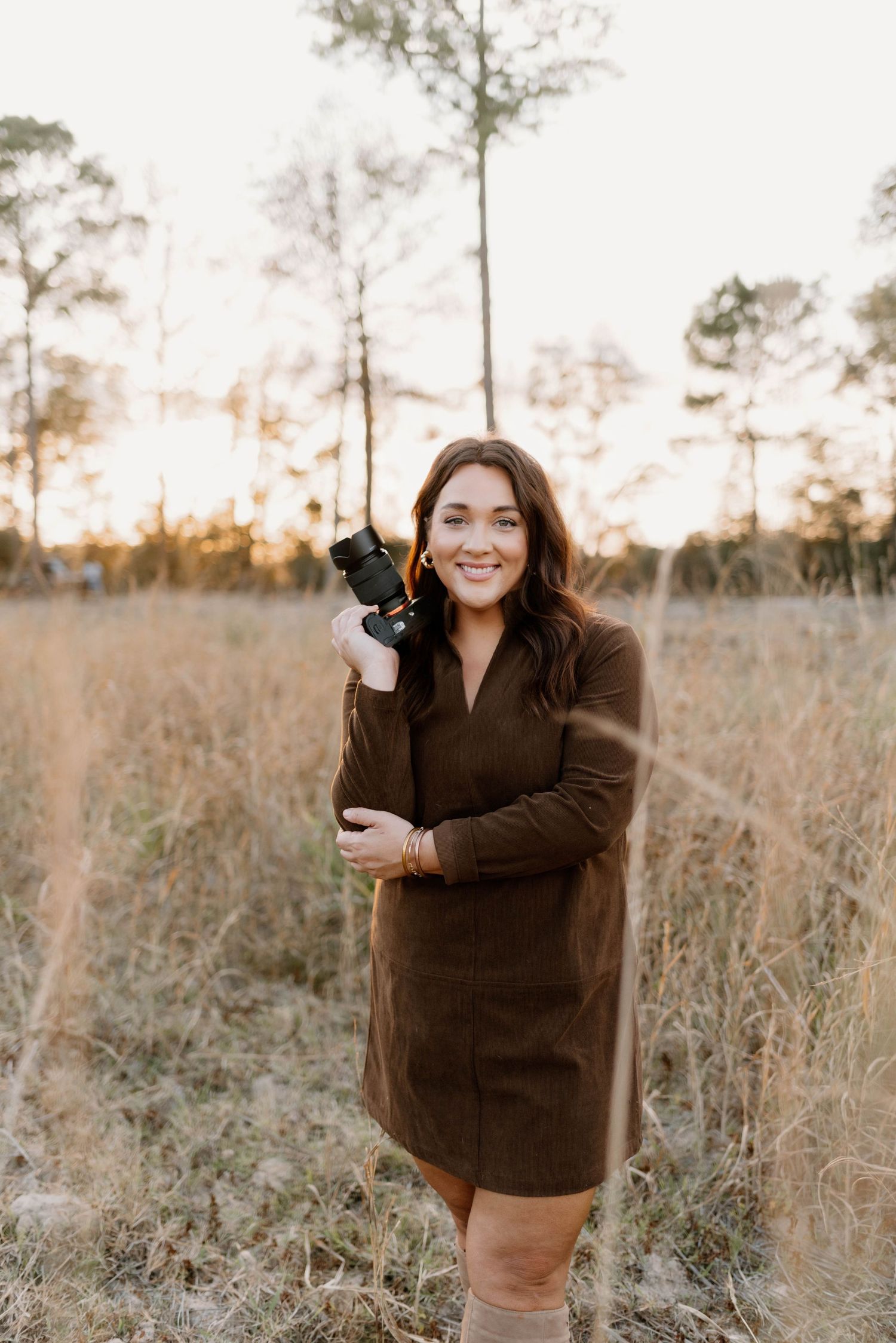 A photographer in a brown sweater dress stands in a golden field at sunset holding a professional camera.