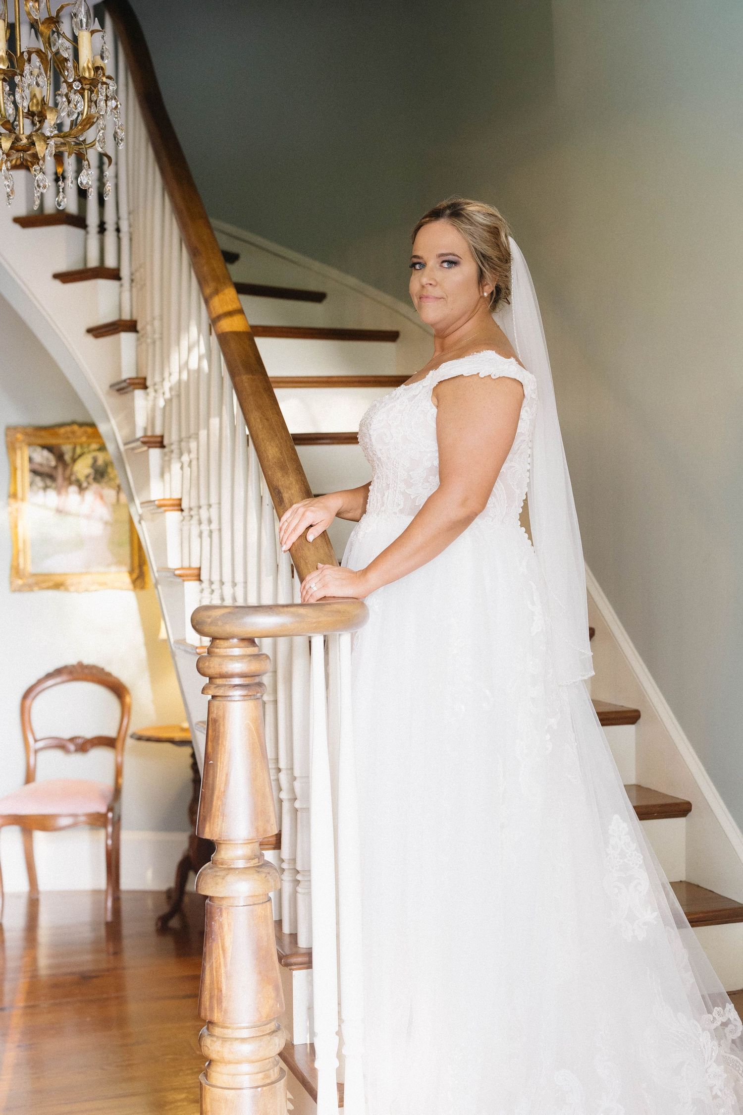 A bride in a white wedding gown poses elegantly on a wooden staircase with white railings in an indoor setting.