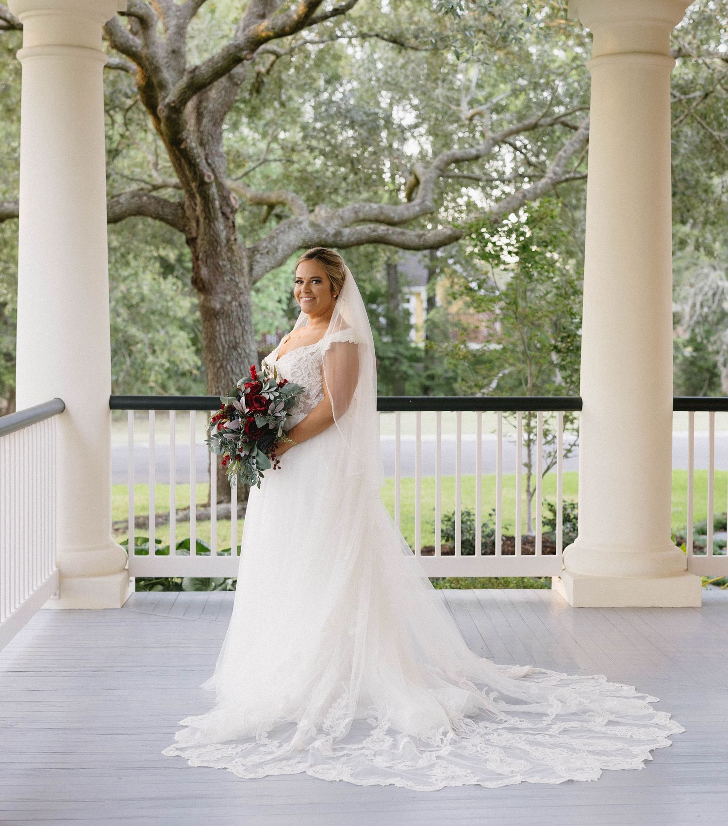 A bride in a flowing white gown with a long train stands gracefully on a columned porch surrounded by trees.