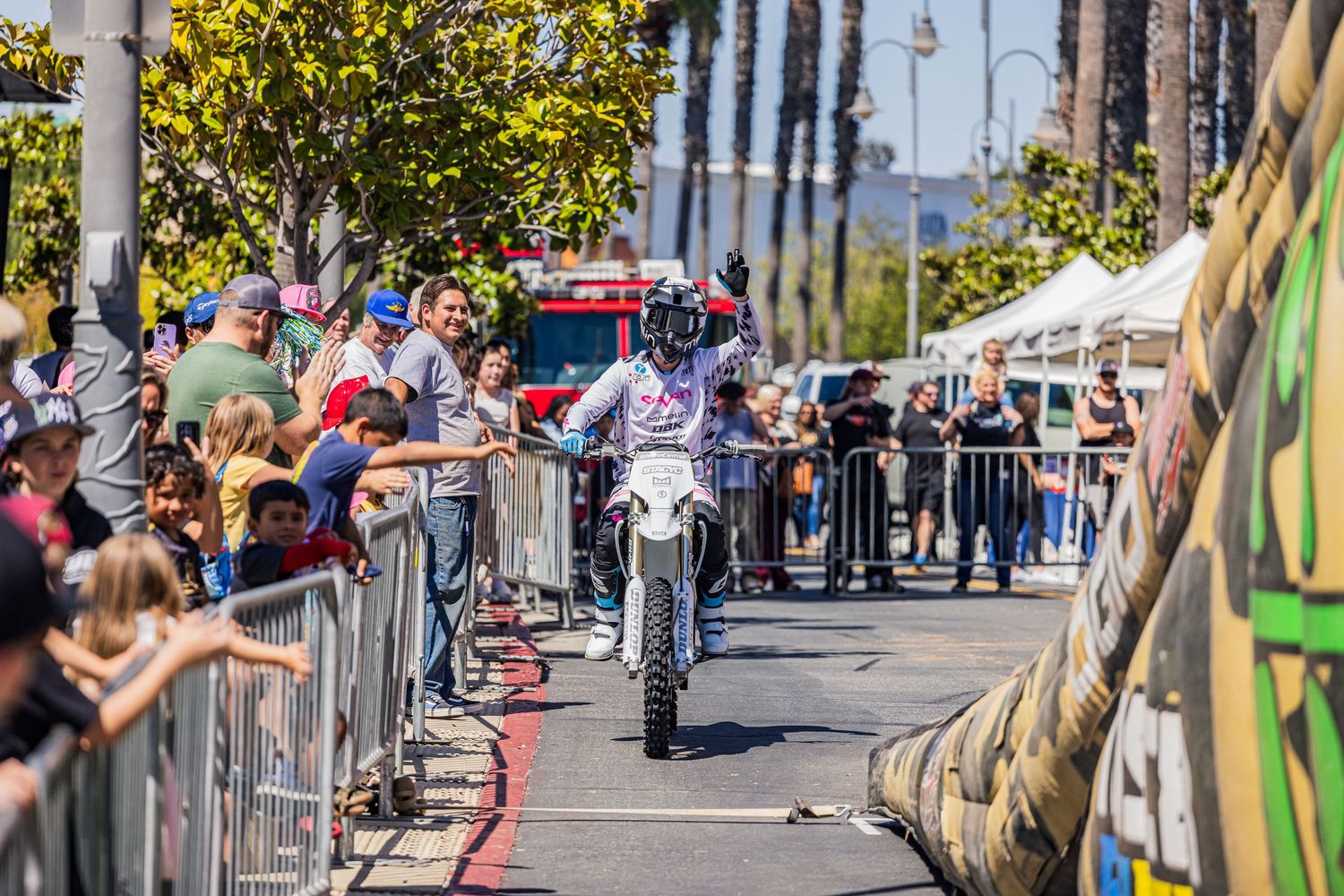 Motorcyclists performing stunts on a sunny street with spectators watching from behind barriers.
