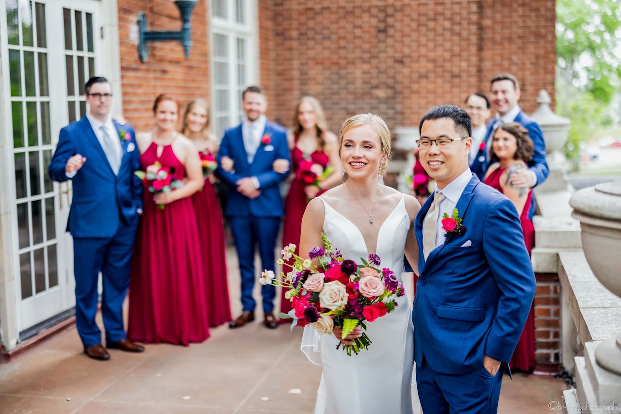Wedding party in navy suits and red dresses gathered outside brick building.