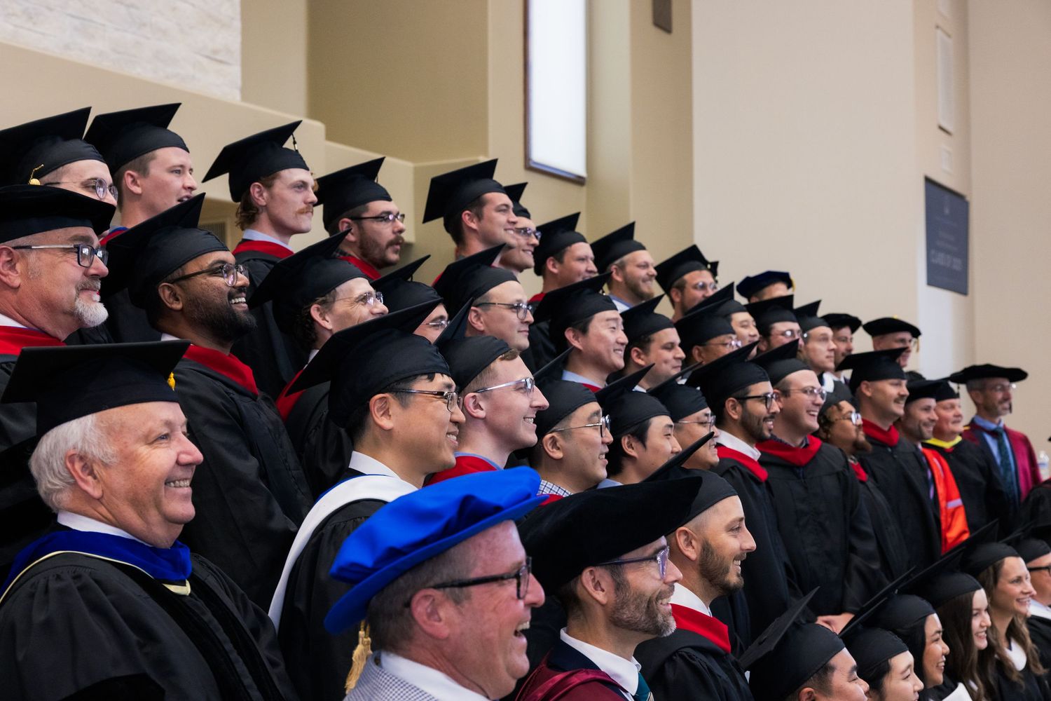 Row of graduates in academic regalia viewing ceremony from elevated seating area.