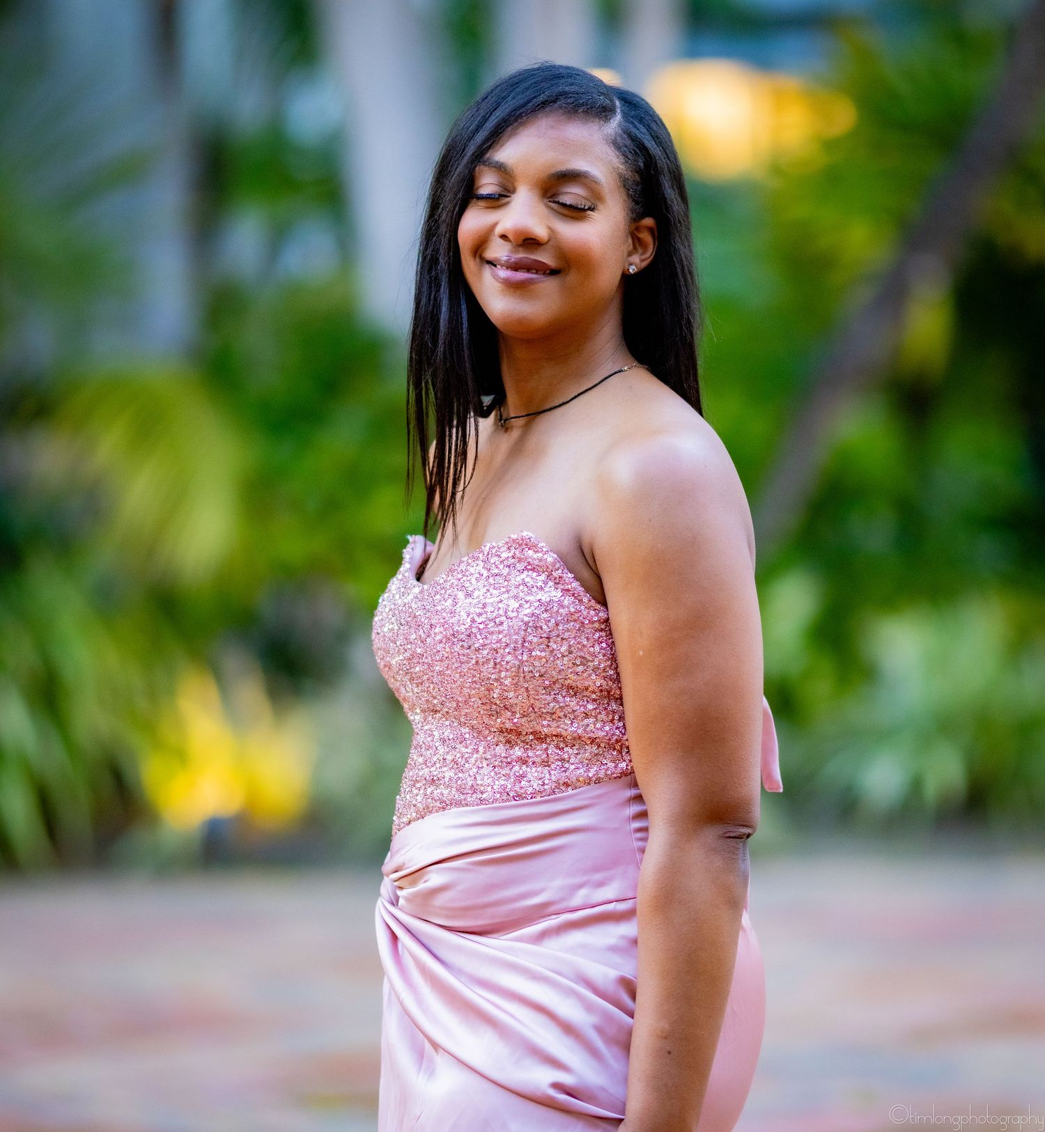 Person in pink sequined formal dress smiles while posing outdoors among tropical foliage.