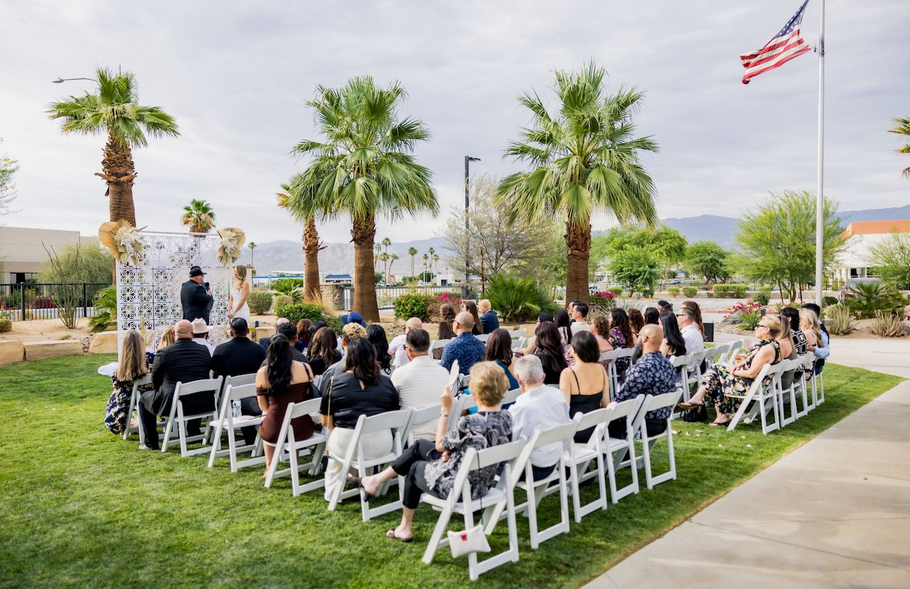 Outdoor wedding ceremony setup with white chairs and palm trees under a bright sky with American flag.