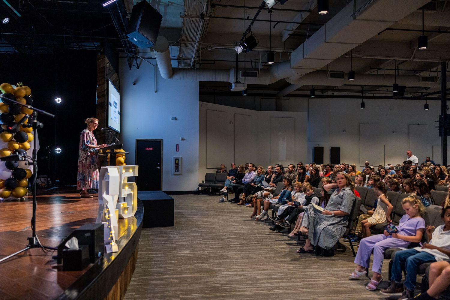 Wide view of audience members seated in rows at a dimly lit indoor conference or presentation.