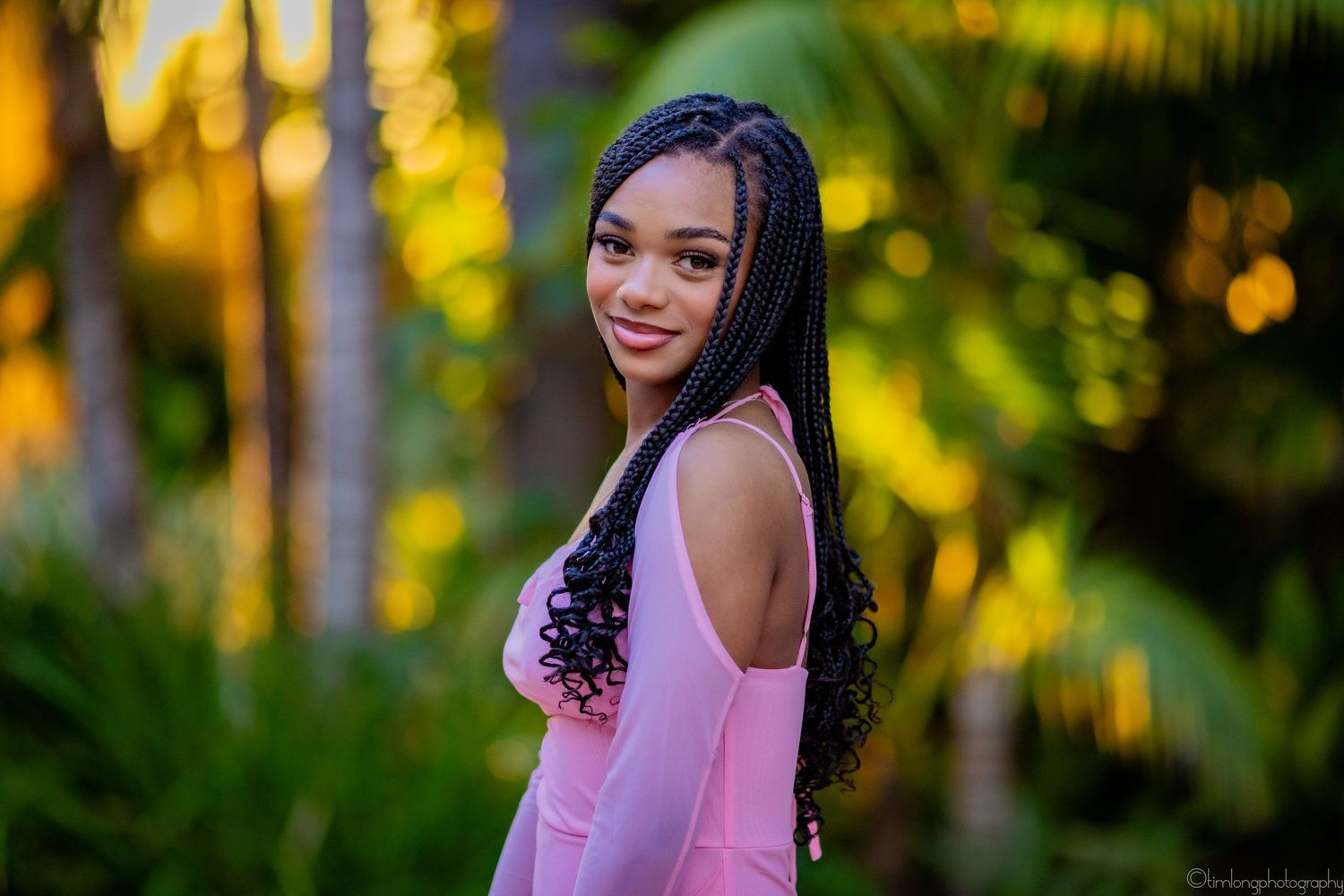 Person in pink off-shoulder dress poses outdoors with palm trees at golden hour.