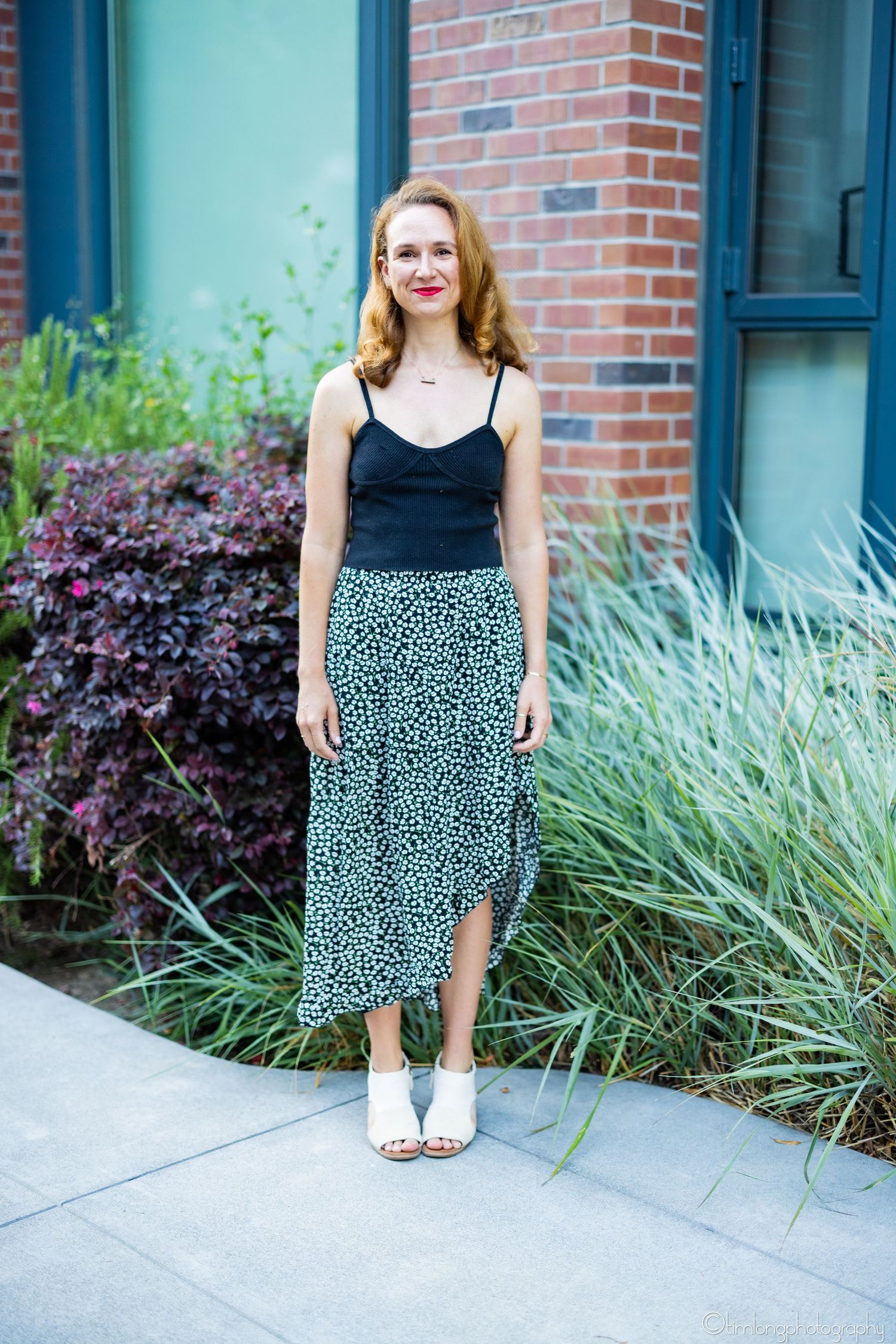 Woman in black top and leopard print skirt stands by brick wall and ornamental grasses.