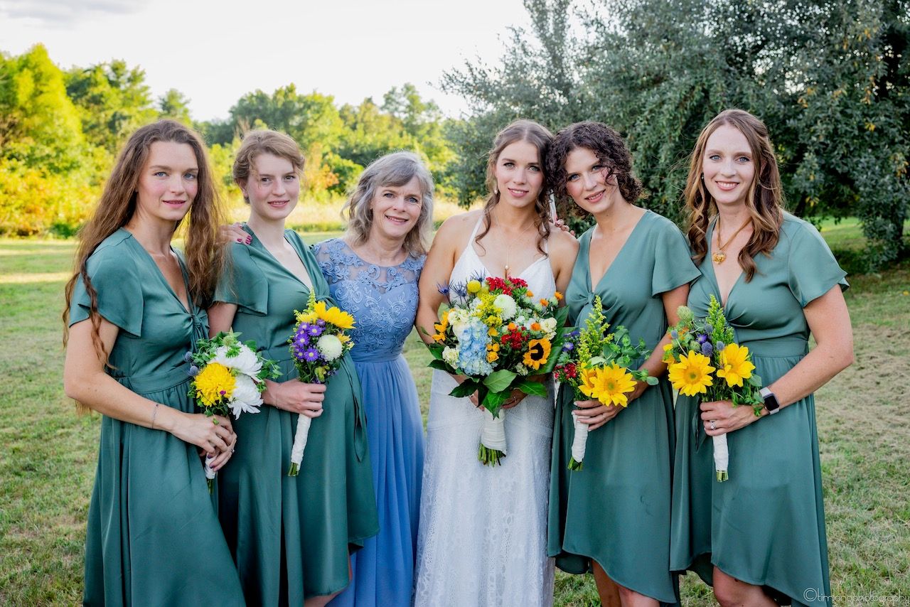 Wedding party in sage green dresses holding vibrant yellow and orange bouquets outdoors.