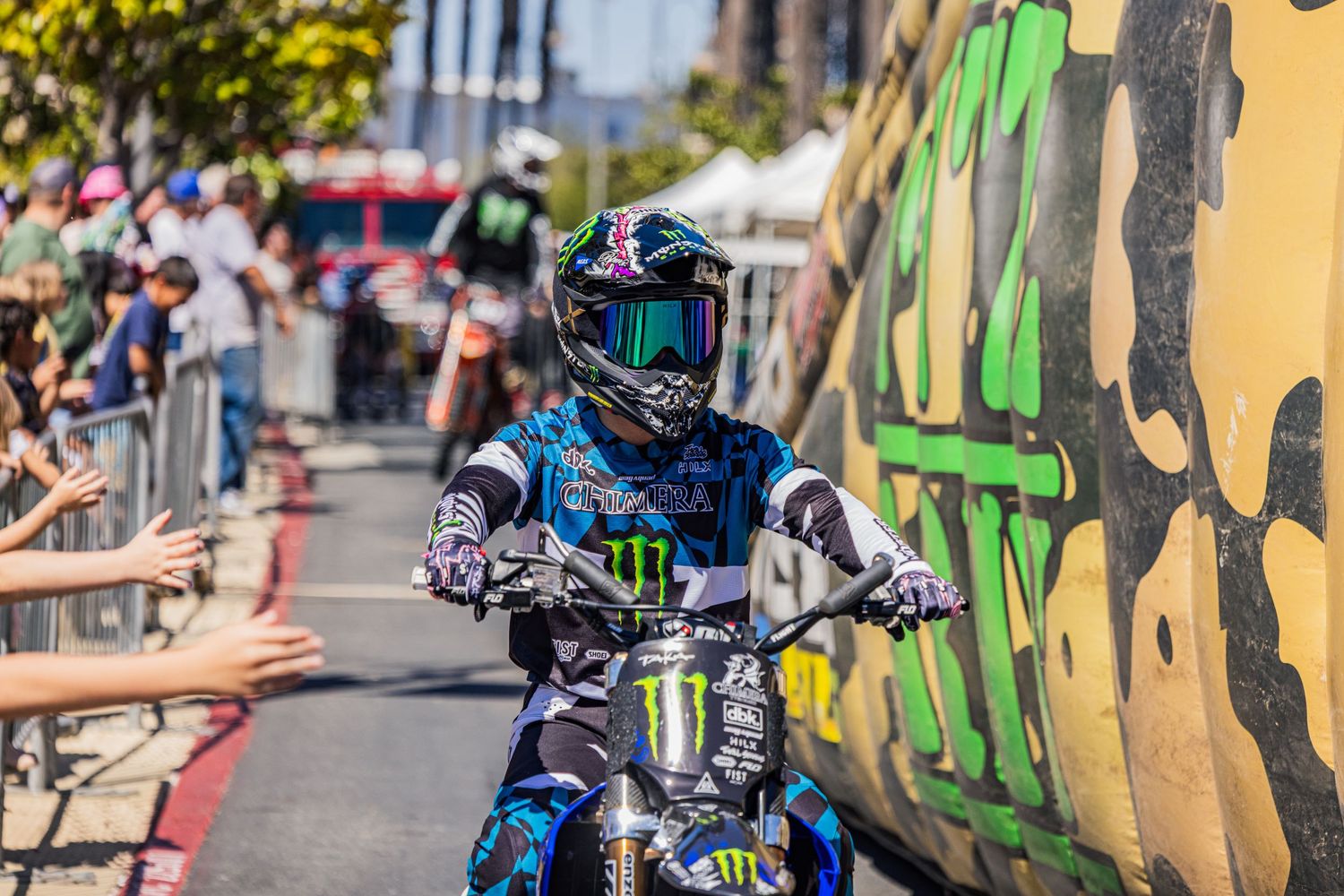 Motorcycle stunt show with riders executing tricks along a barricaded street with cheering crowd.