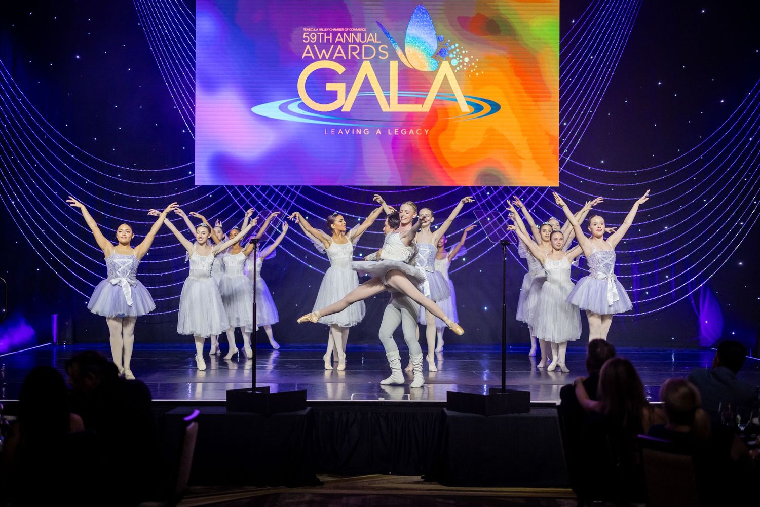 Ballet dancers perform in white tutus and costumes on stage with colorful lighting and curved line backdrop designs.