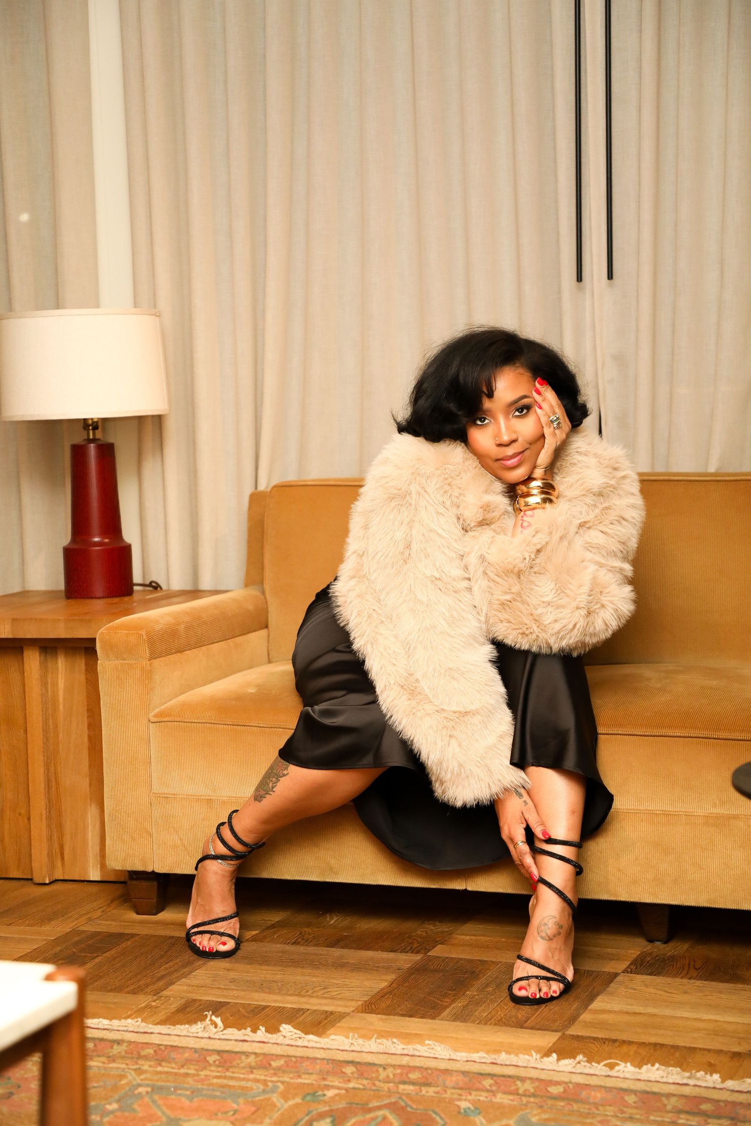 A figure in a black dress and fluffy white fur stole poses dramatically on a hardwood floor near a beige couch.