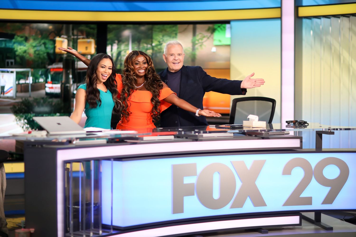 Television news anchors sit behind desk during broadcast at Fox 29 studio as shown in multiple monitor displays.