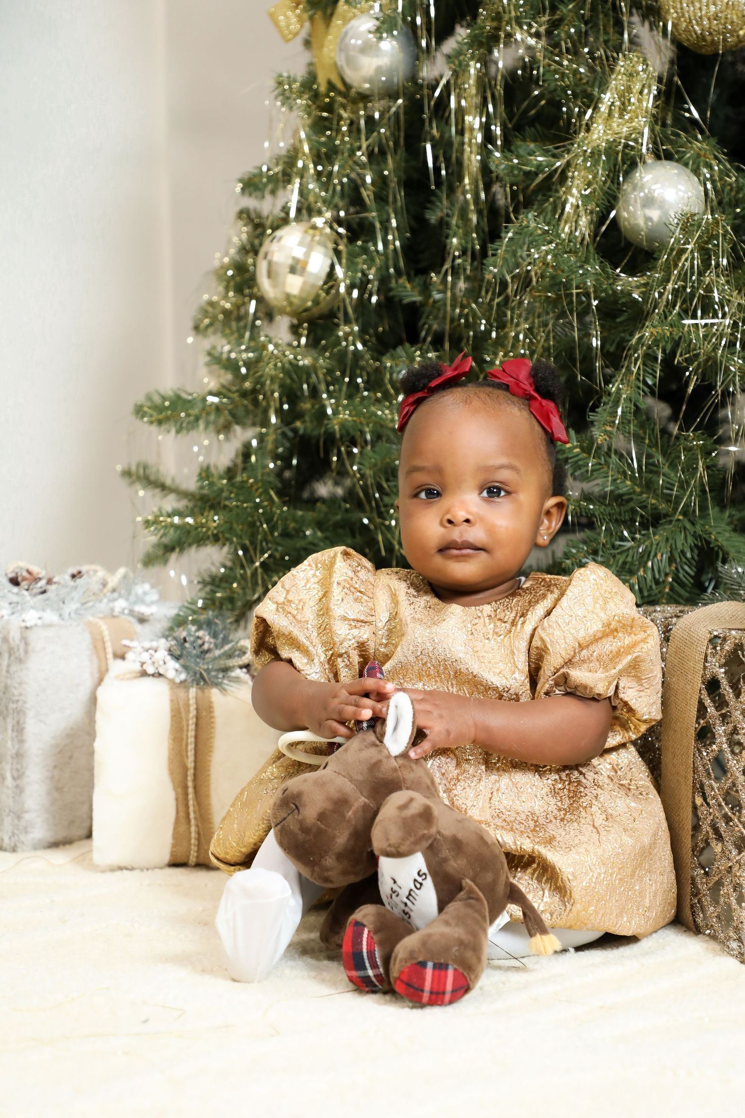 Baby in gold holiday dress sits by Christmas tree with wrapped presents.