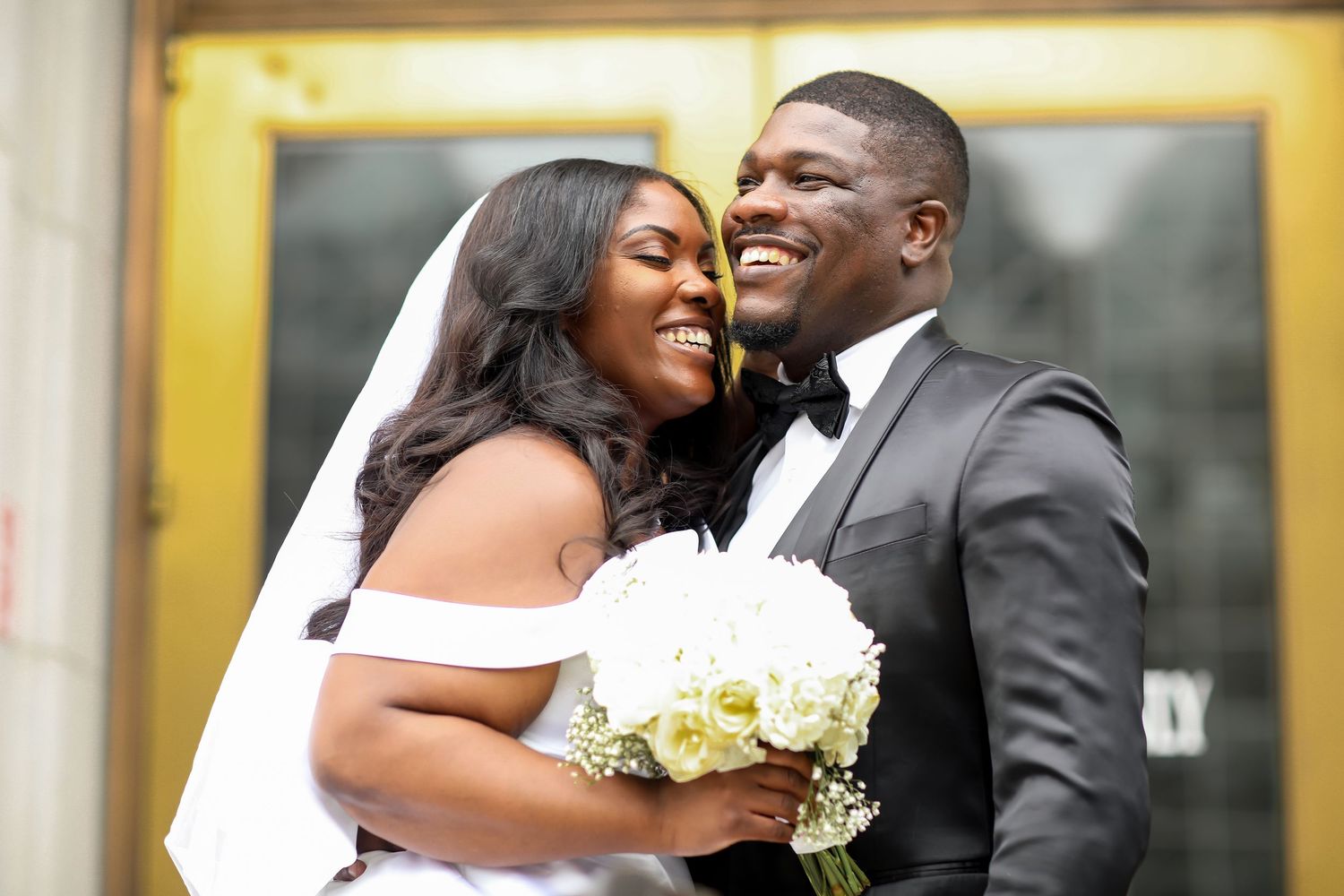 A wedding couple shares intimate moments in front of a yellow-lit building.