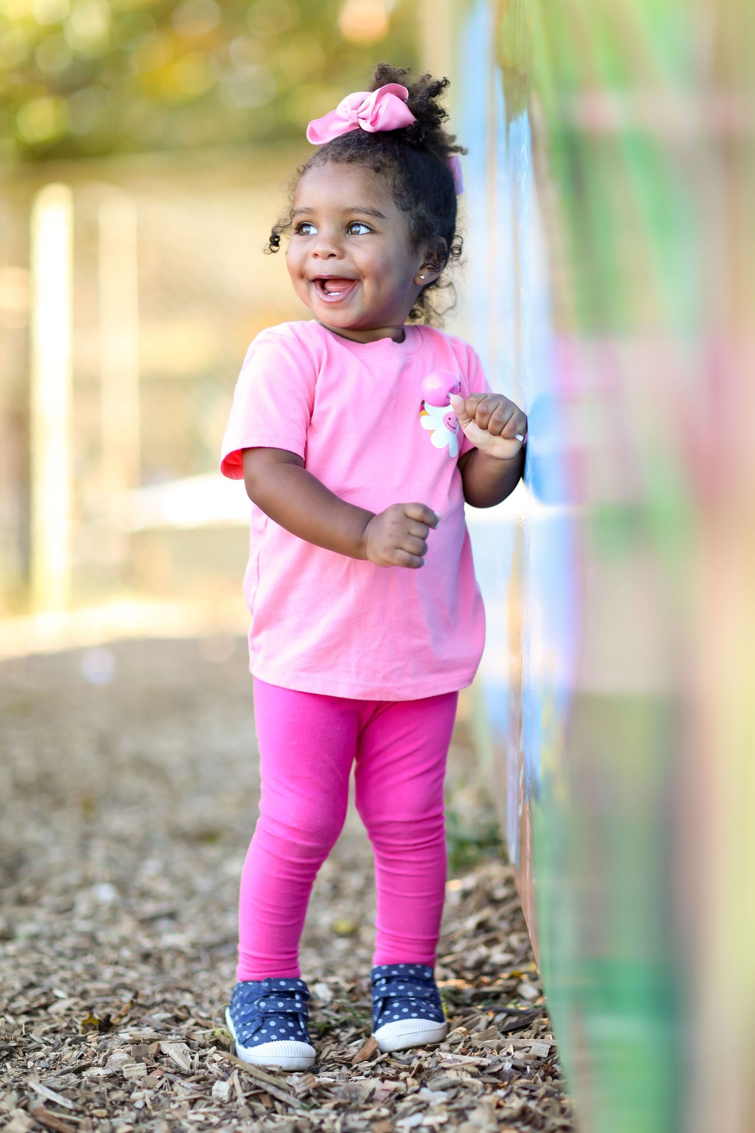 A small child in bright pink clothing and polka dot shoes explores outdoors in dappled sunlight.
