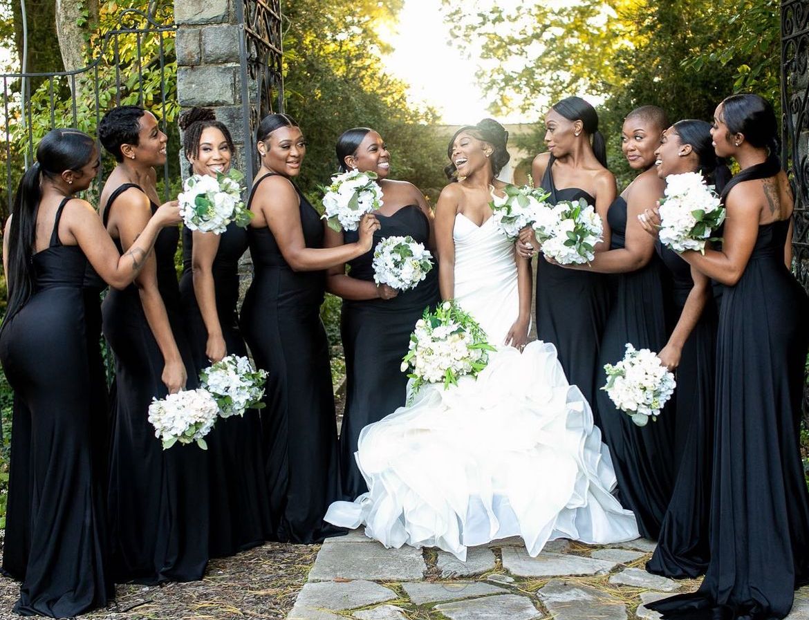 Bridesmaids in black dresses pose with the bride in a white gown during an outdoor wedding.