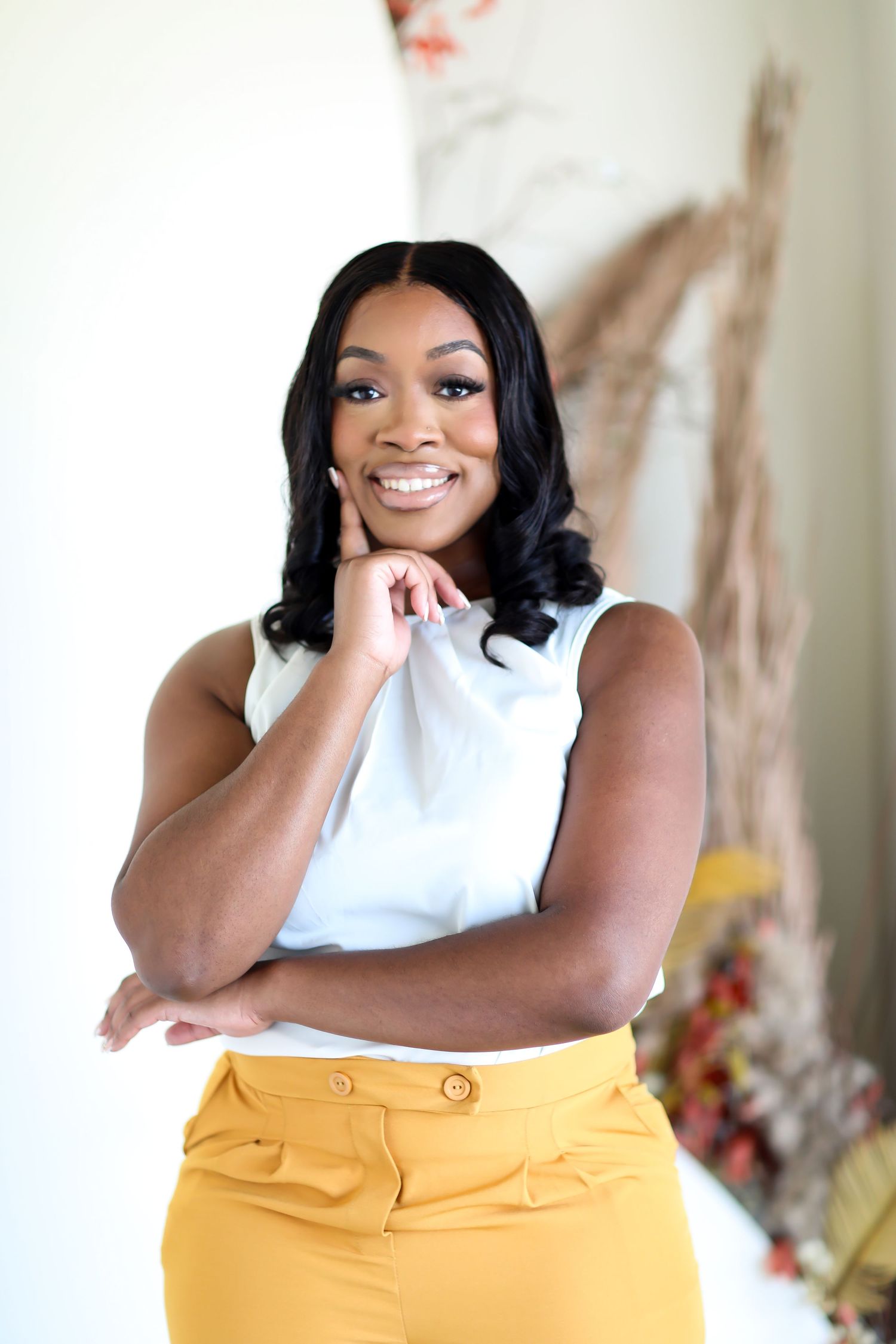 Woman in white sleeveless top and yellow pants poses with hand under chin against light background.